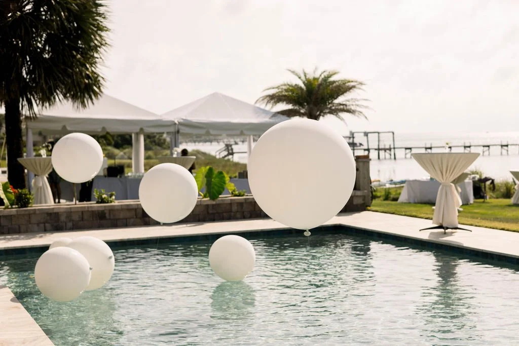 Poolside party setup with floating white balloons, outdoor tables, and canopy tents near a body of water with palm trees and a pier.
