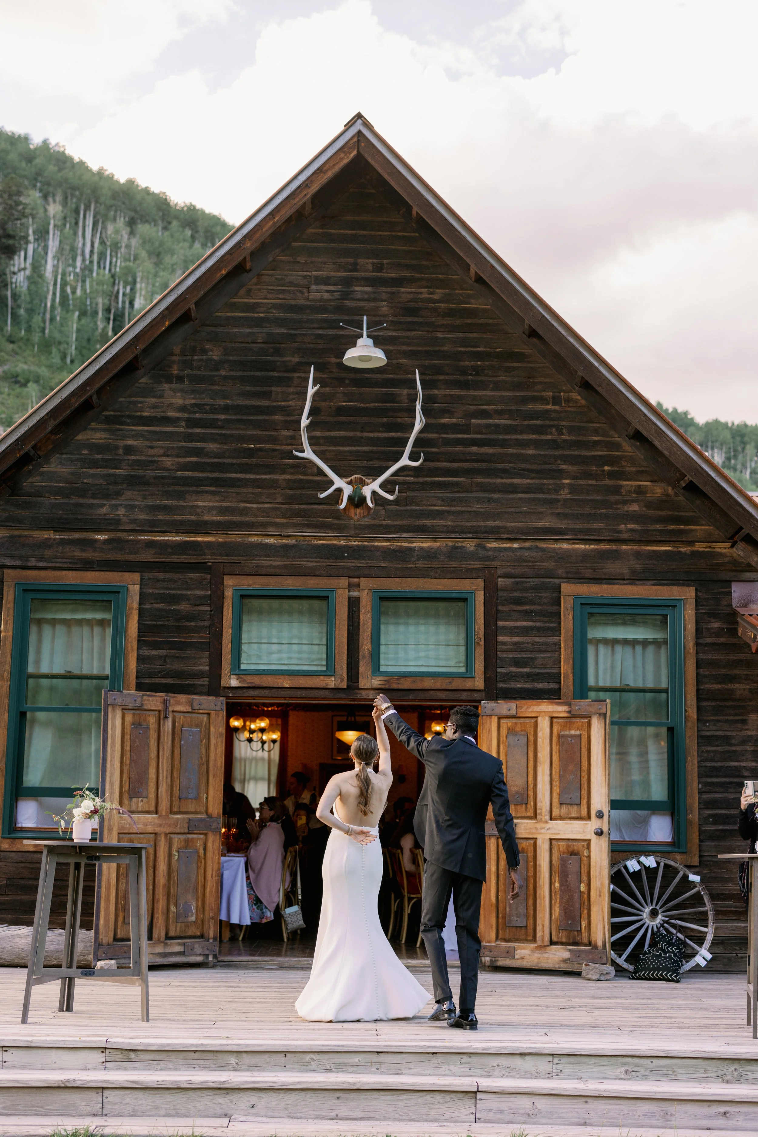 A bride and groom dancing in front of a rustic wooden barn with open doors, antler chandelier, and mountain scenery in the background.