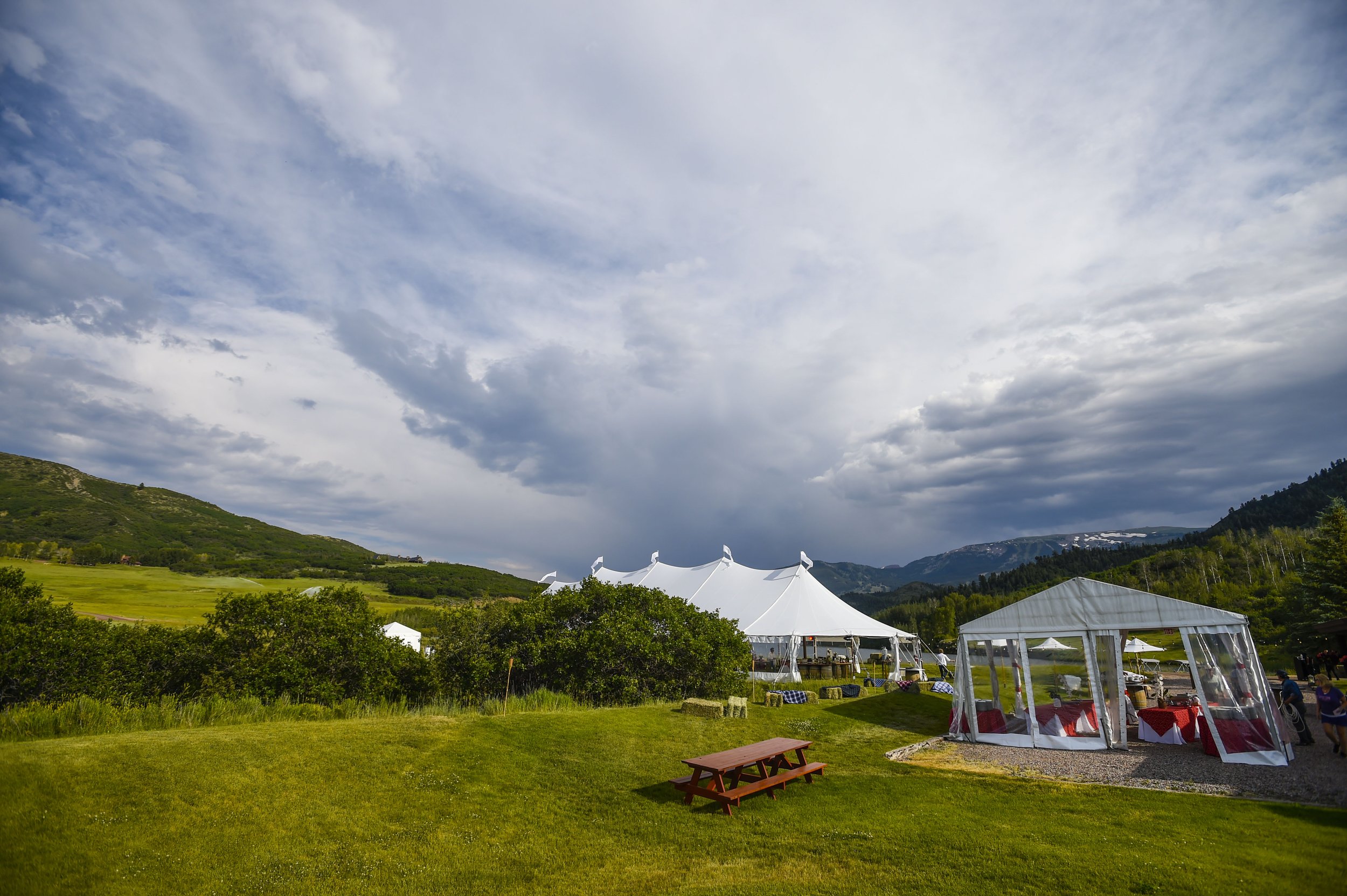 Outdoor event setup in a green field with white tents, picnic tables, and tables with red and white tablecloths, surrounded by mountains and trees under a partly cloudy sky.