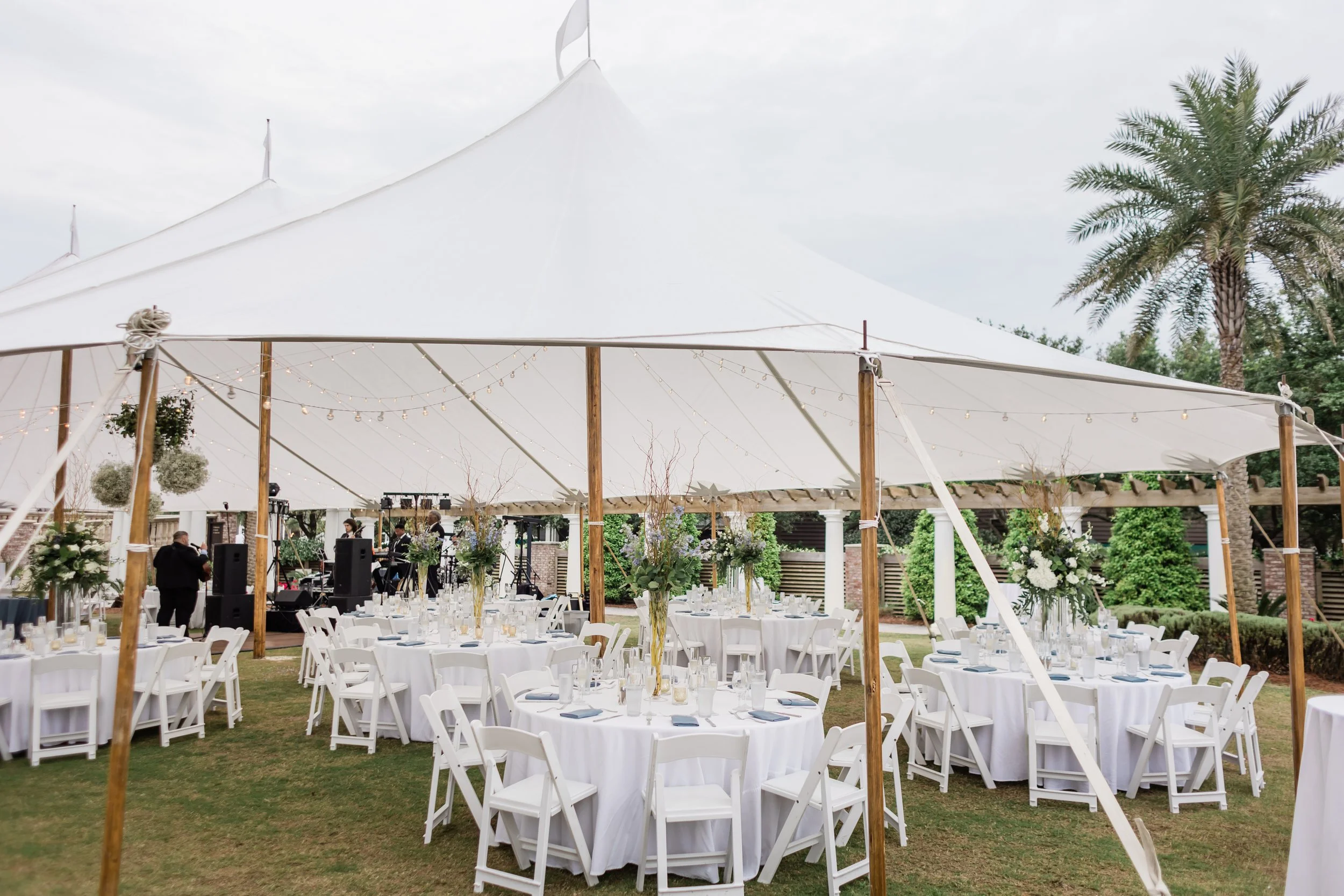 Outdoor event setup with round tables covered in white tablecloths, decorated with tall floral centerpieces, under a large white tent with string lights, surrounded by greenery and palm trees.