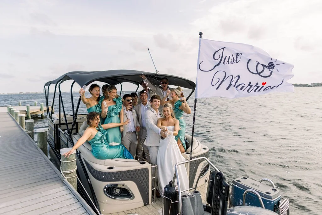 A group of people in formal wedding attire celebrating on a boat with a 'Just Married' flag.