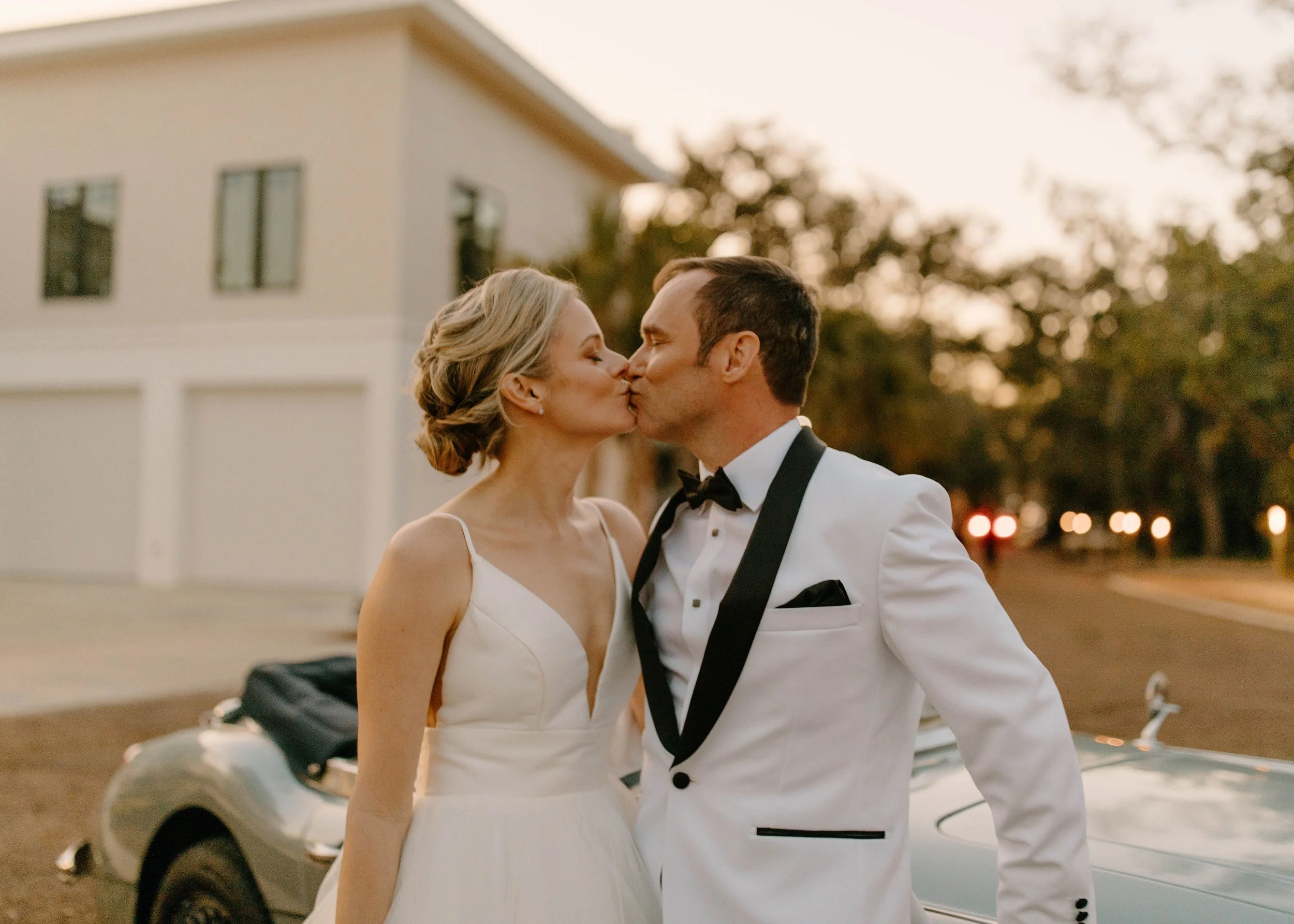 A bride and groom in wedding attire sharing a kiss outdoors at sunset.