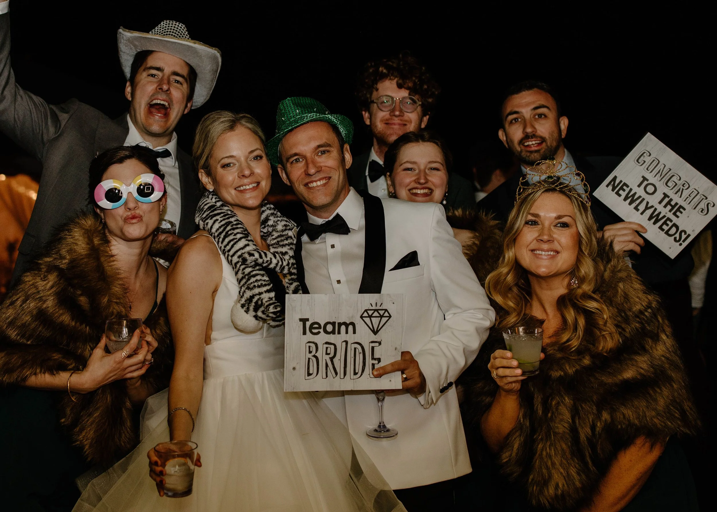 Group of people celebrating at a wedding reception wearing fun costumes and holding drinks. The bride and groom are in the center, with the bride holding a sign that says 'Team Bride' and the groom holding a glass of champagne. Others are wearing wig