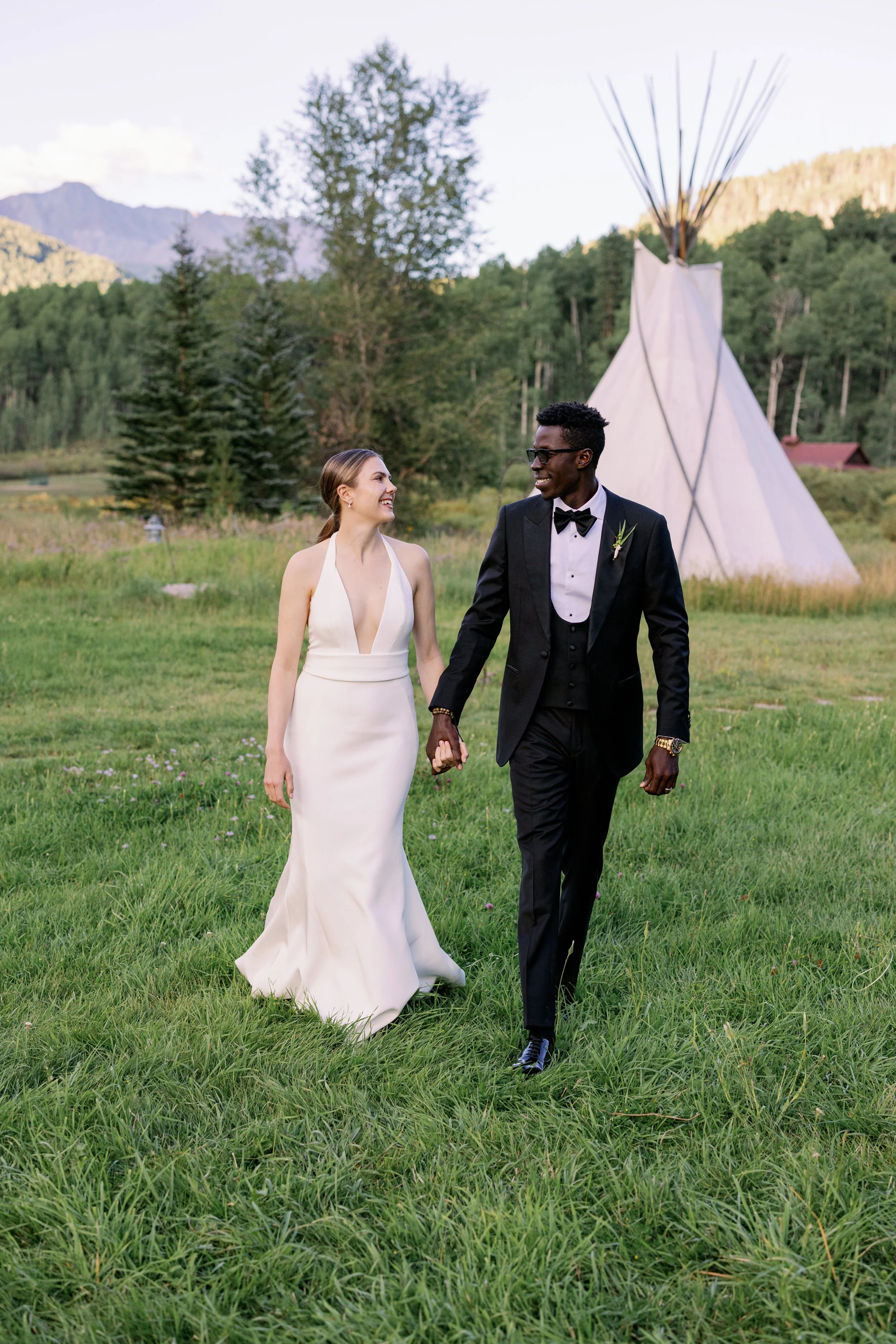 Bride and groom walking hand in hand on a grassy field during their wedding, with a tipi in the background and mountains and trees behind them.