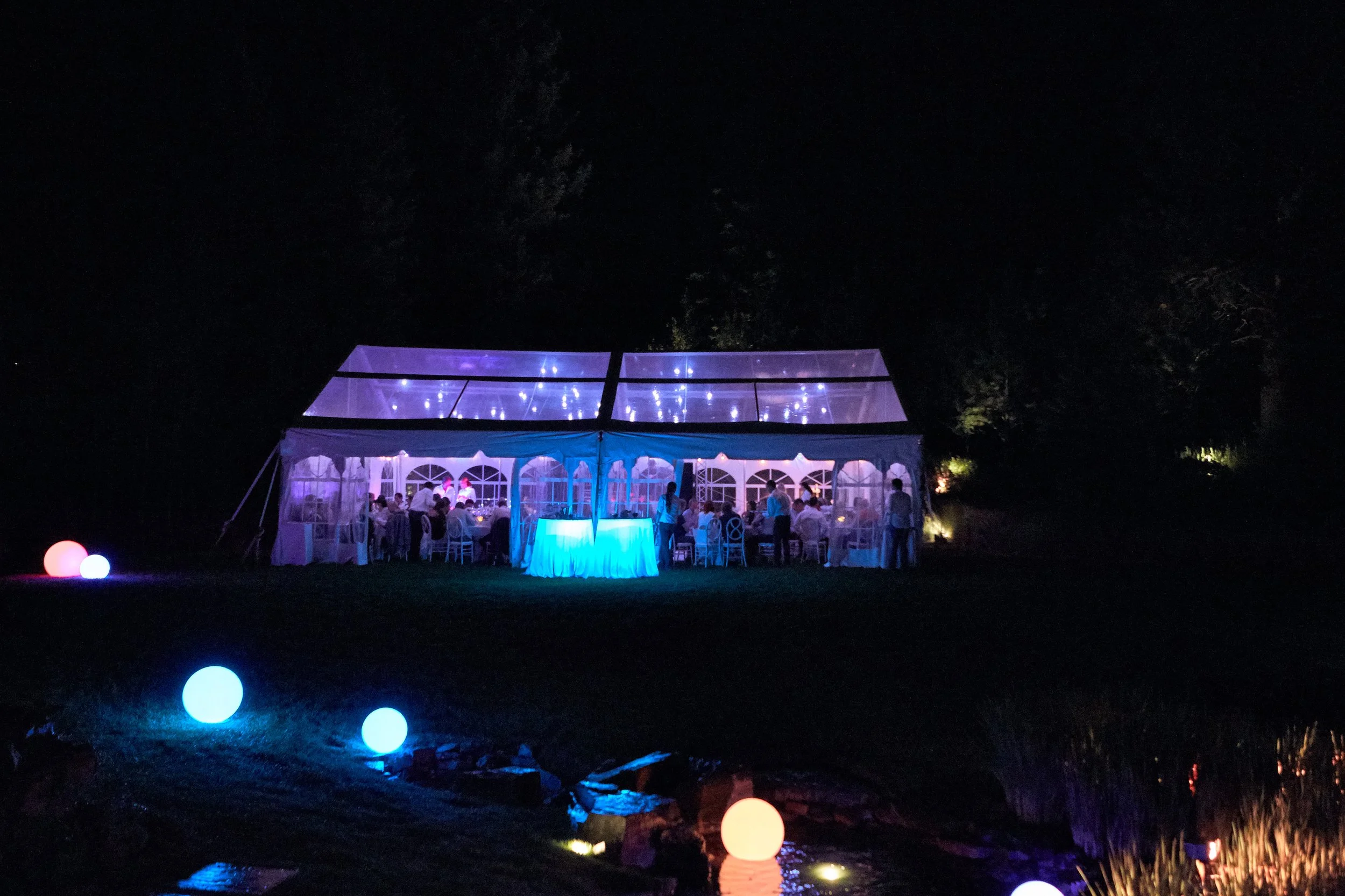Nighttime outdoor event with a large, illuminated tent decorated with string lights. Several people are gathered inside and around the tent, which has transparent panels. Colorful illuminated spheres are on the ground in the foreground, and the scene