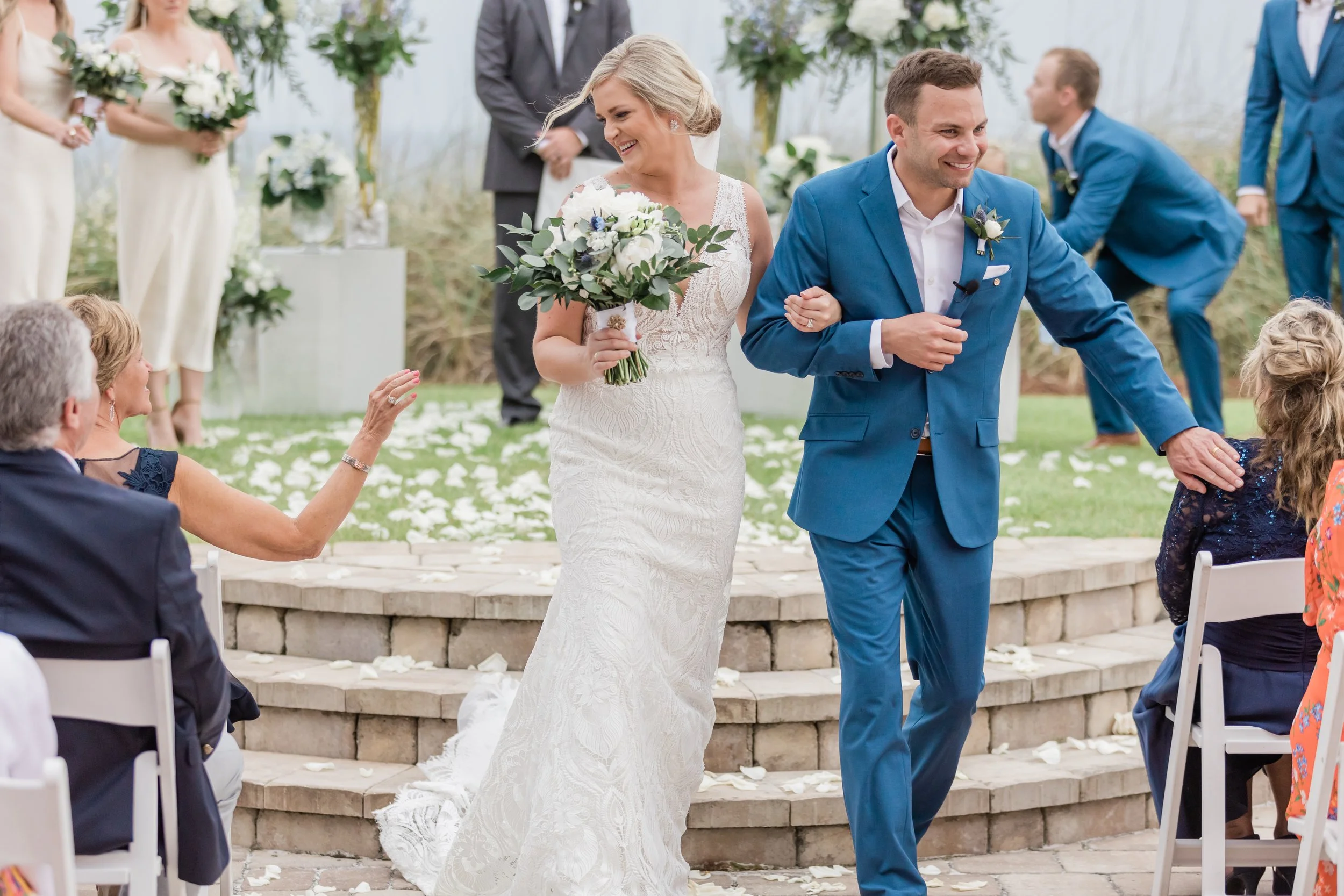 A bride and groom walk down the aisle at their outdoor wedding ceremony, smiling and holding hands, with wedding guests seated nearby.