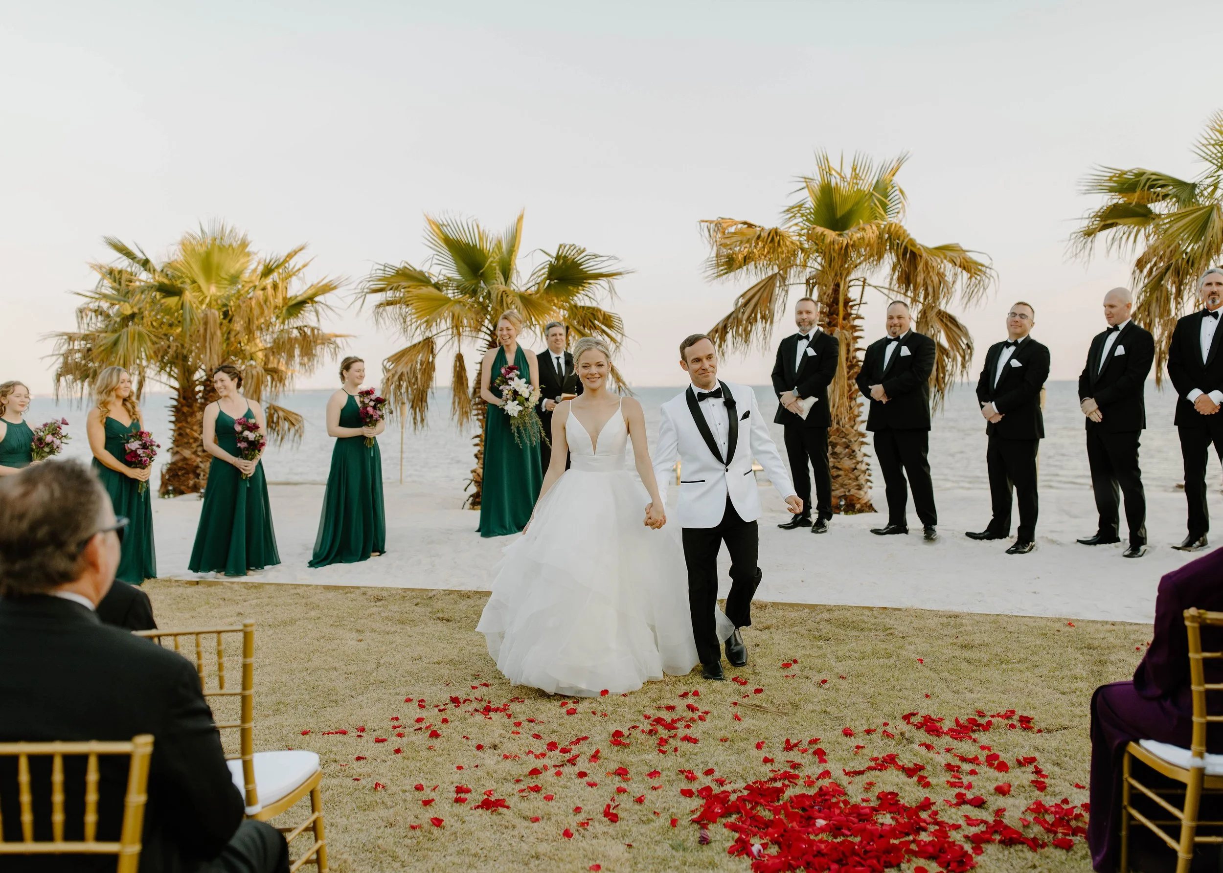 A wedding ceremony takes place on a beach with the bride and groom walking hand in hand, surrounded by bridesmaids in green dresses holding bouquets and groomsmen in tuxedos, with palm trees and the ocean in the background.