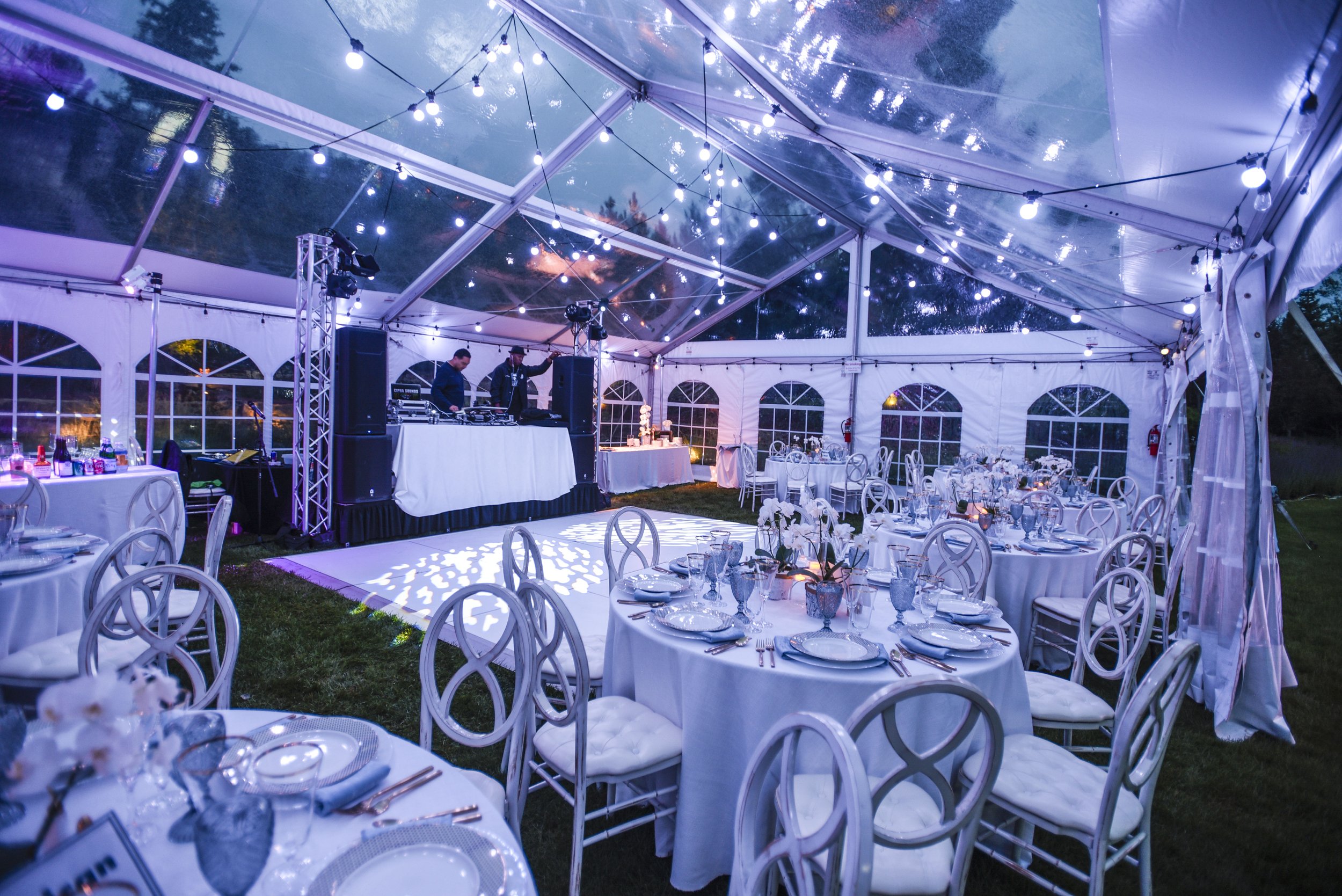 Elegant outdoor event tent with decorated tables, stage with DJs, and string lights at dusk.