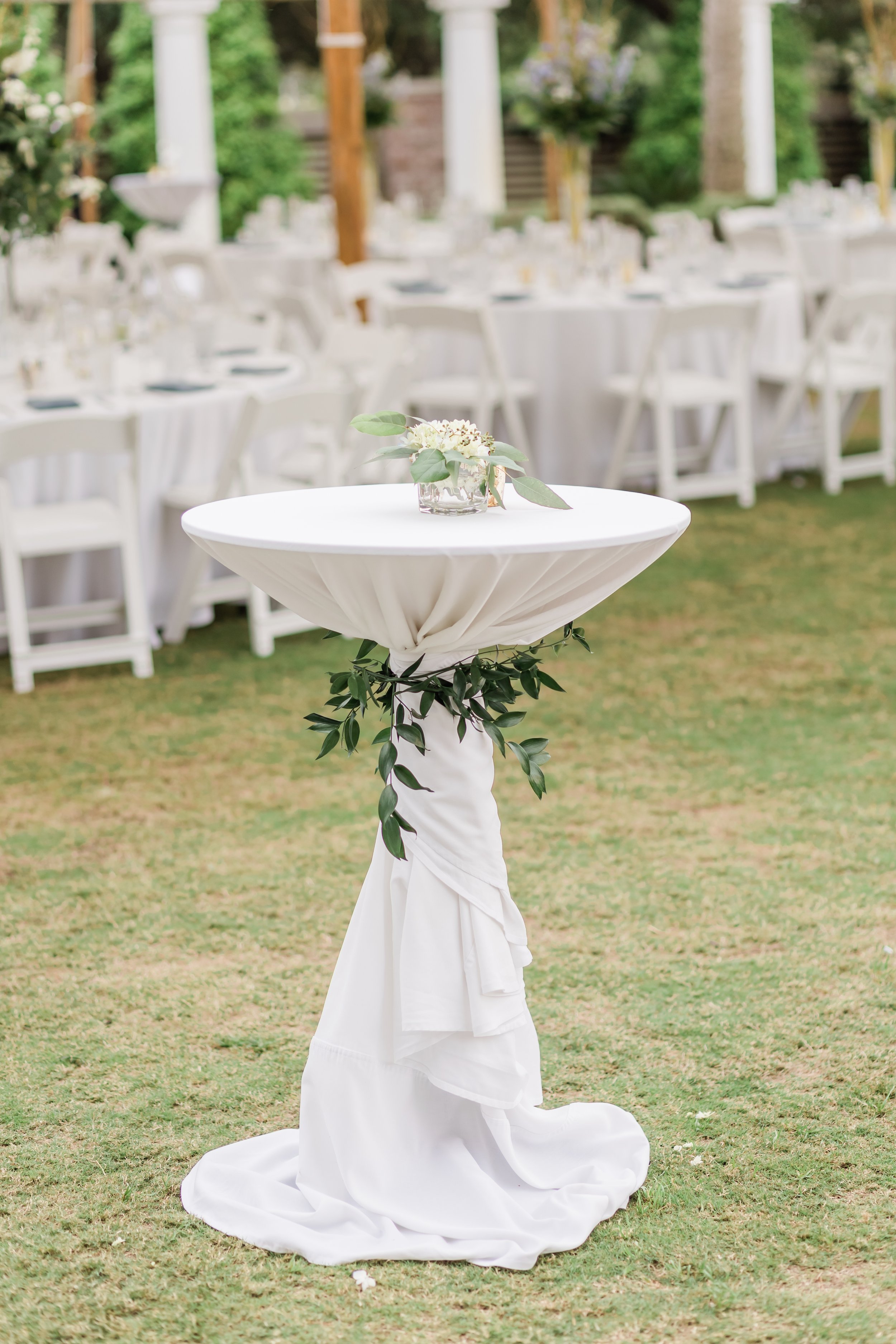 A decorated outdoor wedding reception area with a round, white, fabric-covered table with green foliage accents, featuring a small floral arrangement in a glass container; white chairs and tables with similar decorations are in the background on gras