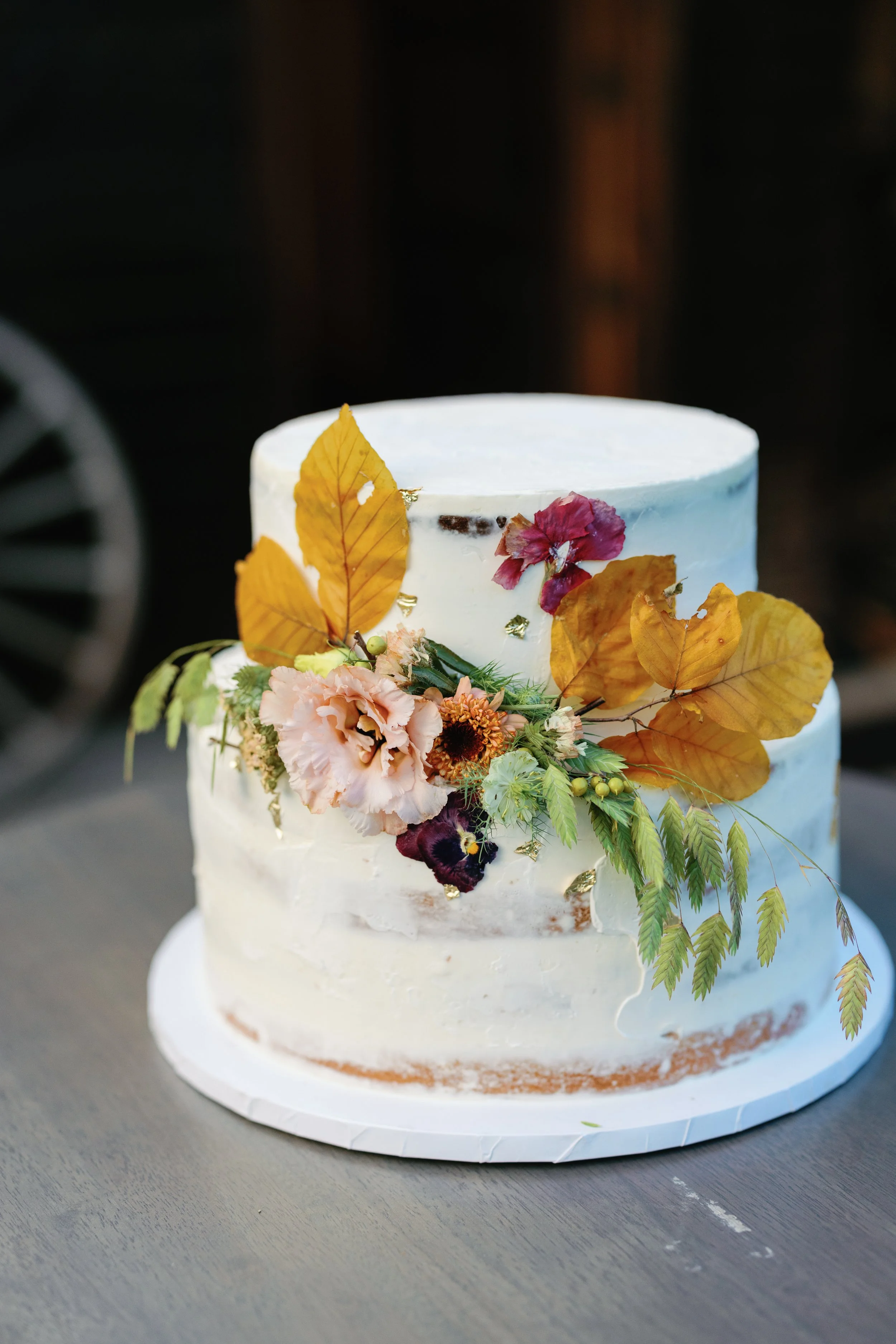 Two-tier white wedding cake decorated with fall foliage and flowers on a gray table.
