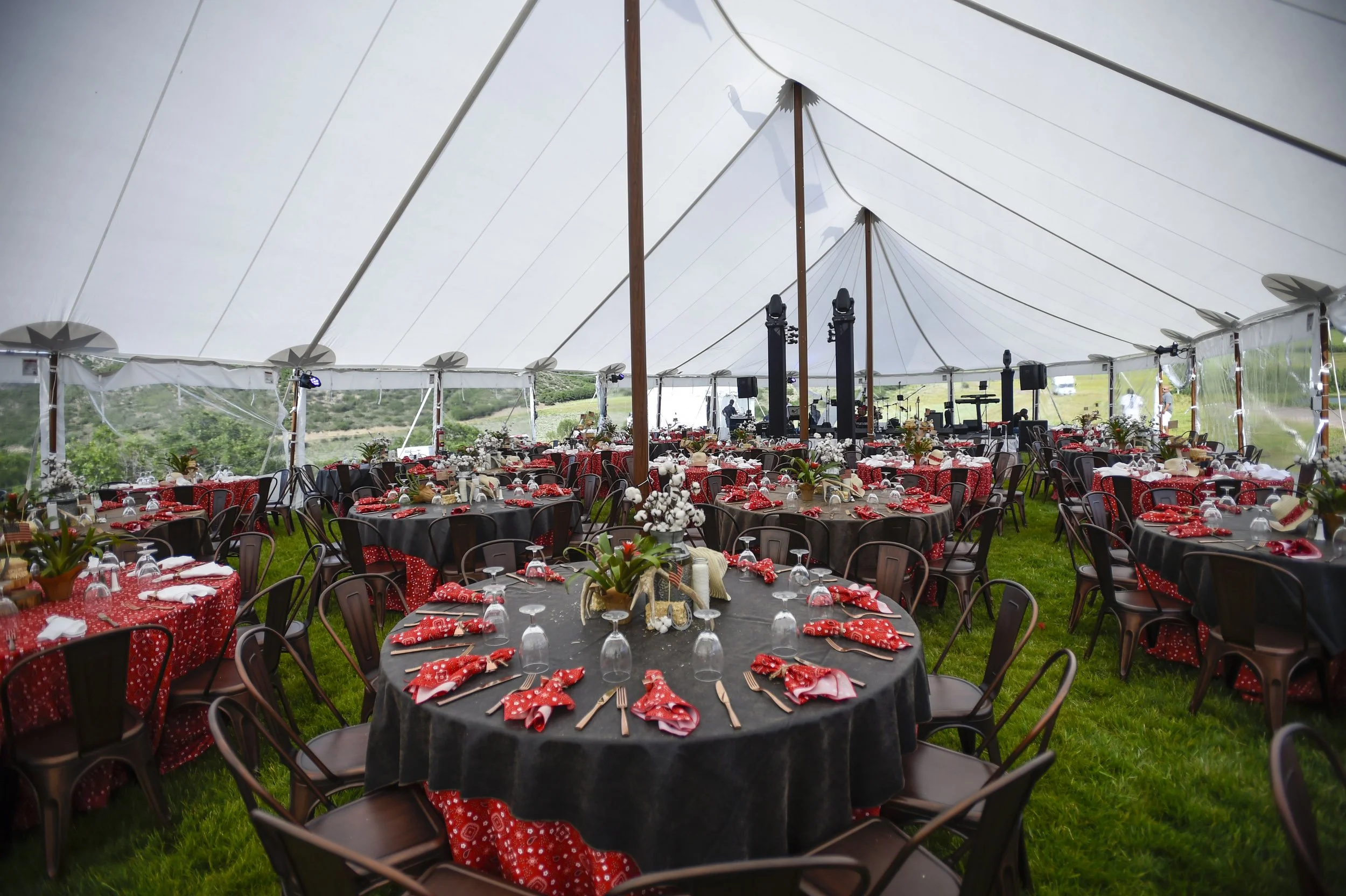 Under a large white event tent, round tables are set for a celebration with black tablecloths, red and white cloth napkins, glassware, and floral centerpieces. Metal chairs are arranged around the tables on green grass, with a stage and musical instr