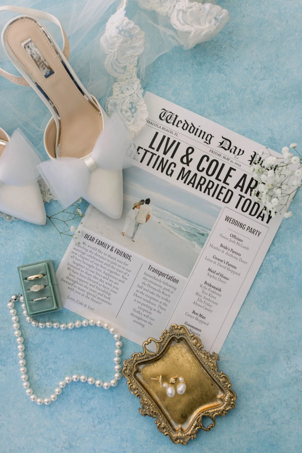 A wedding invitation surrounded by white high-heeled shoes with bows, pearl jewelry, a gold tray with pearl earrings, and decorative flowers on a blue surface.