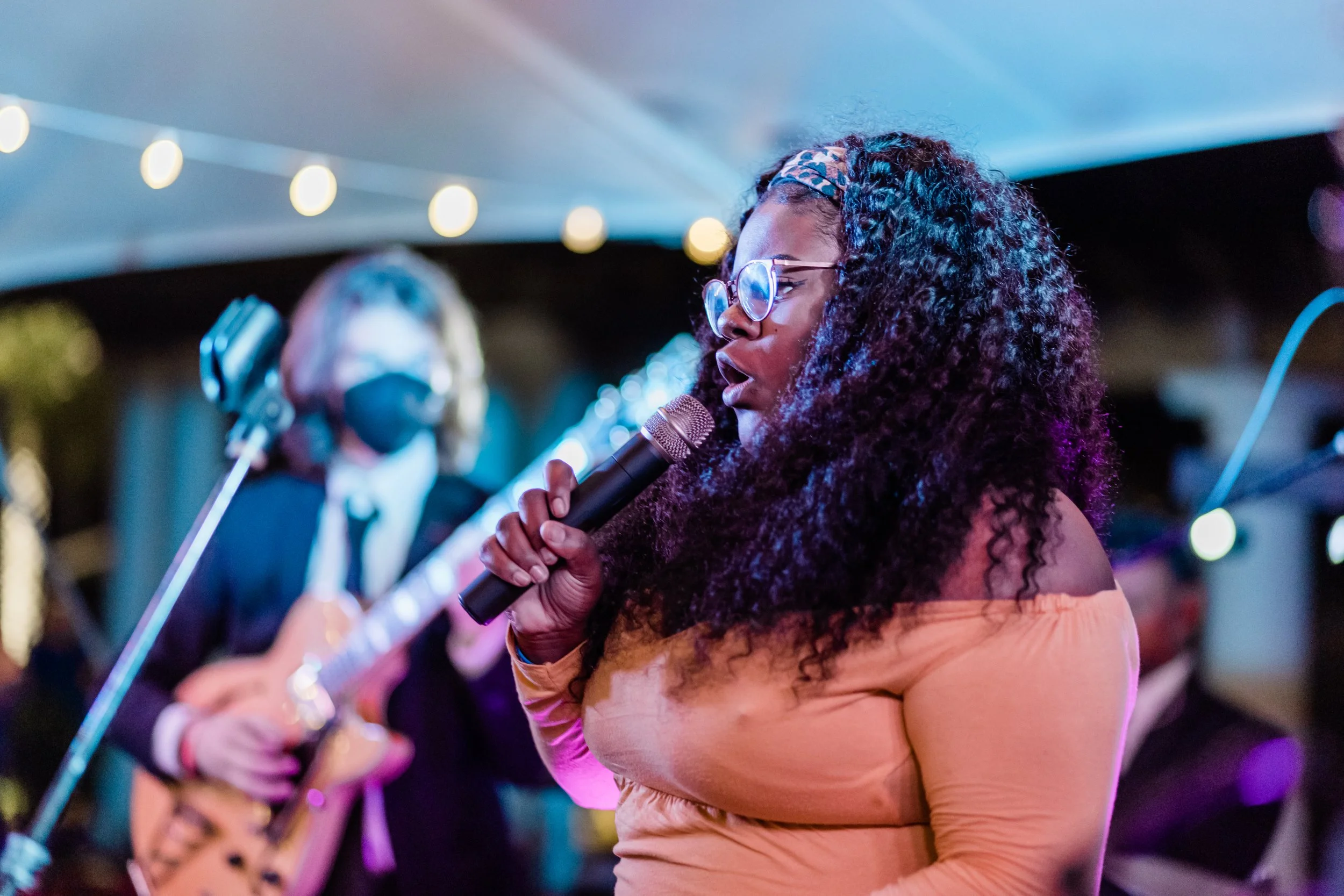 A woman singing into a microphone at an outdoor event with a guitarist playing in the background.