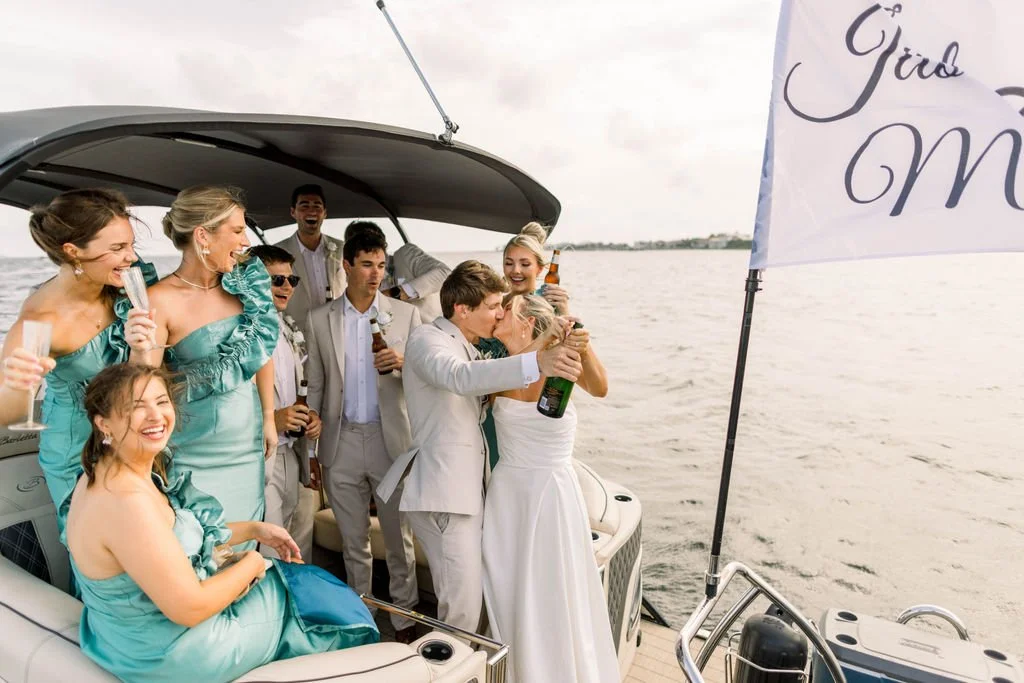 A wedding celebration on a boat with a bride and groom kissing, surrounded by friends in formal attire, holding drinks, and holding a 'Girl Mom' flag, near a body of water.