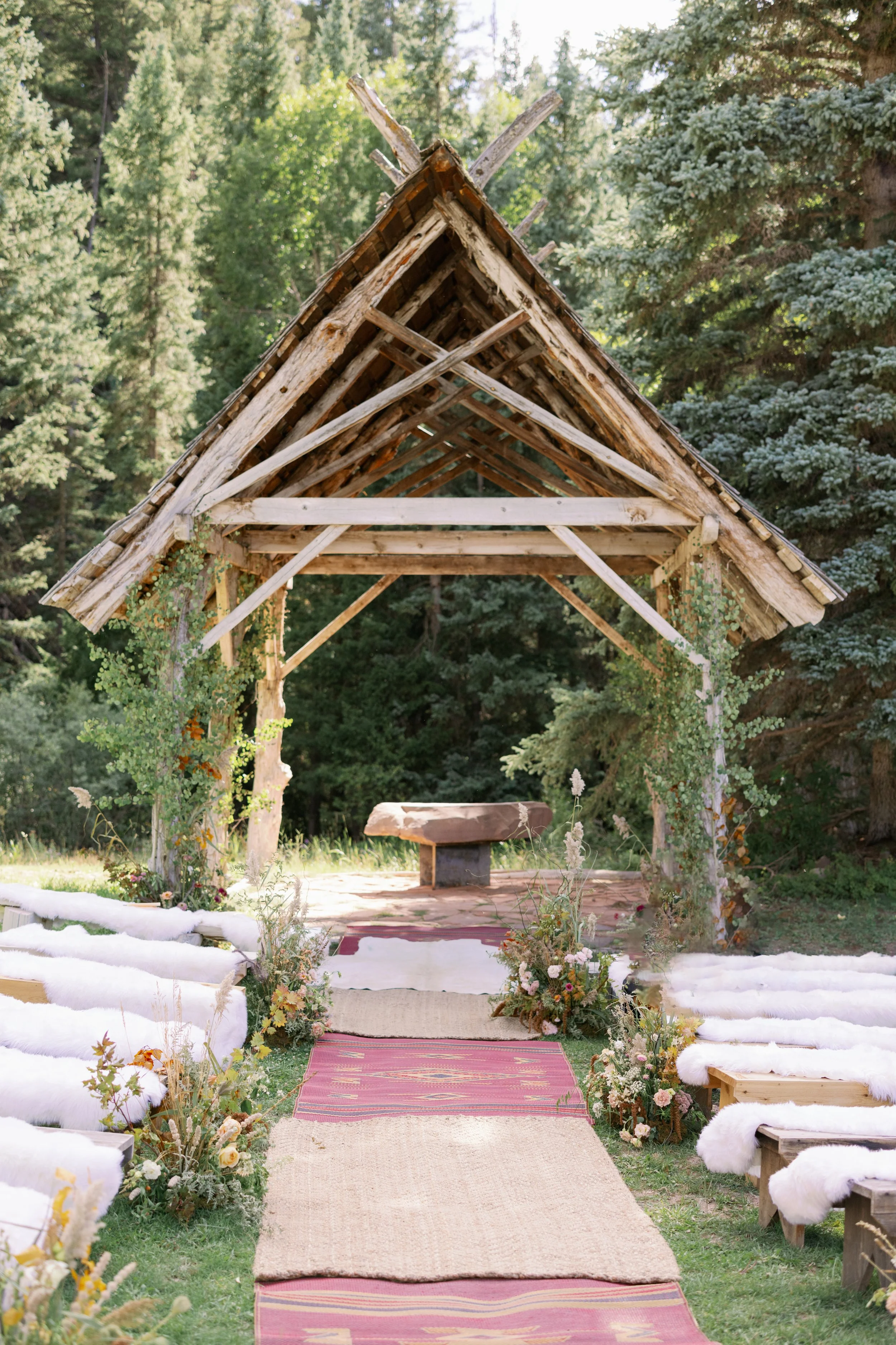 Outdoor rustic wedding altar with a wooden triangular arch, benches draped with white fabric, floral arrangements on either side, and a stone bench at the altar, set in a forested area.
