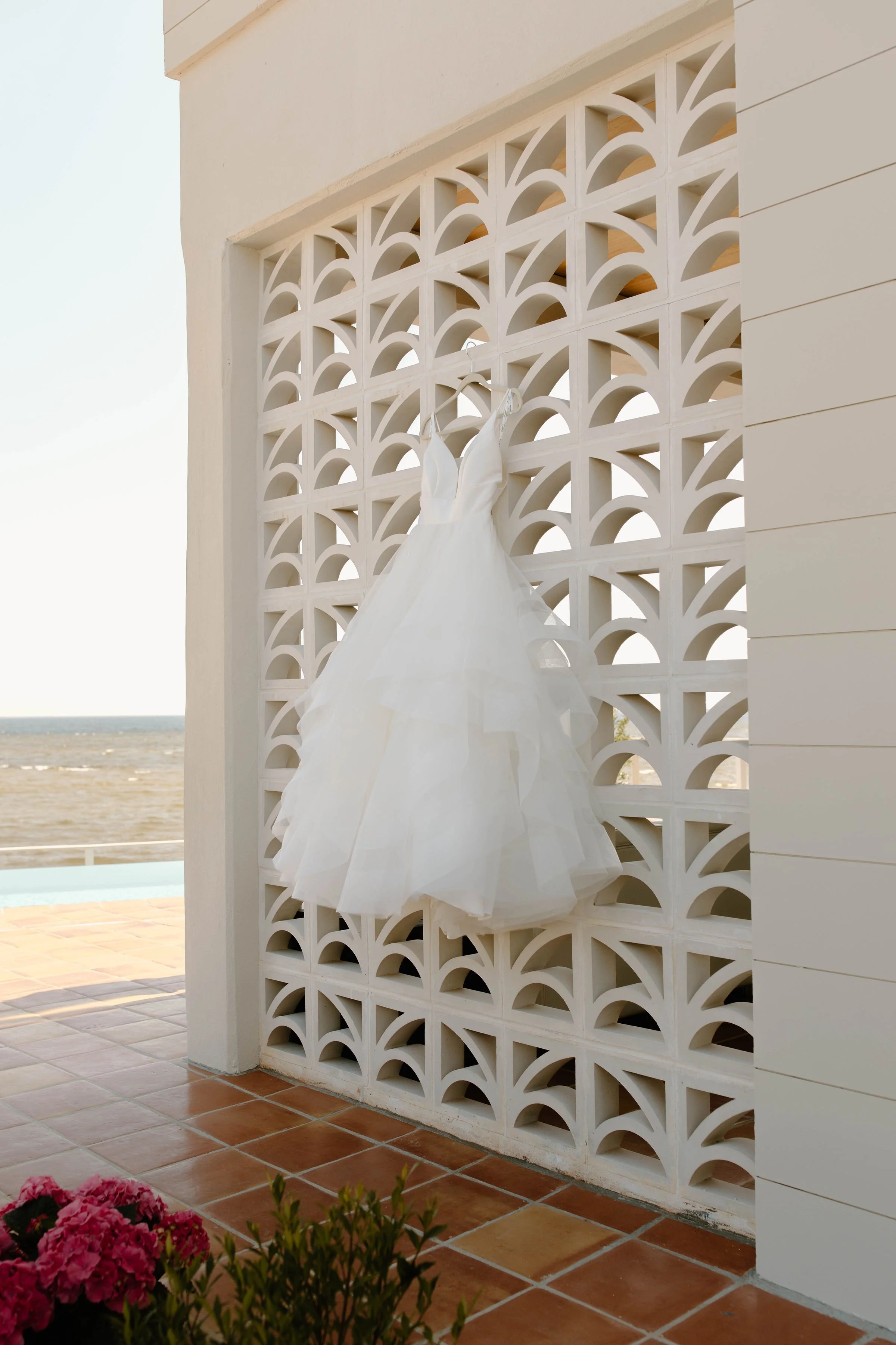A white wedding dress hangs on a decorative white wall with a pattern of geometric cutouts. The scene is set outdoors with a view of a sandy beach and the ocean in the background. Potted pink flowers are visible in the lower corner.