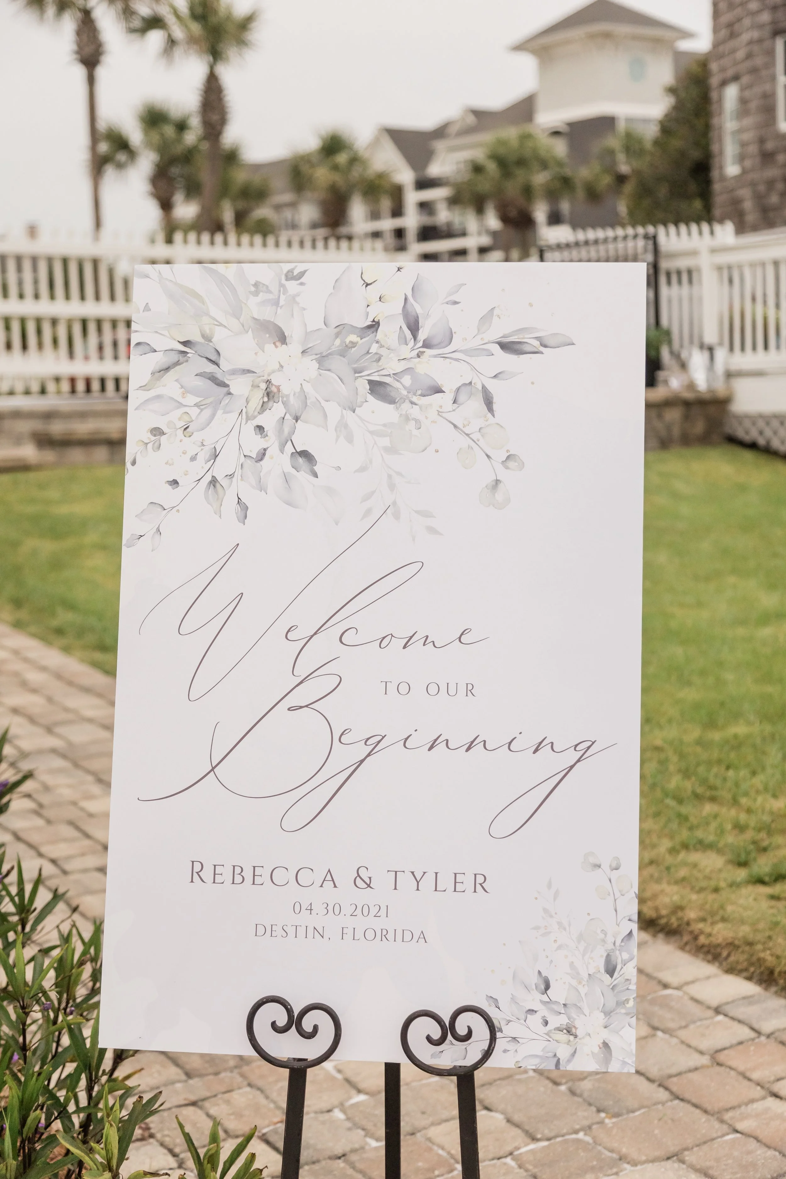 Wedding welcome sign with floral design and elegant script, reading 'Welcome to our beginning, Rebecca & Tyler, 04.30.2021, Destin, Florida' in an outdoor setting.
