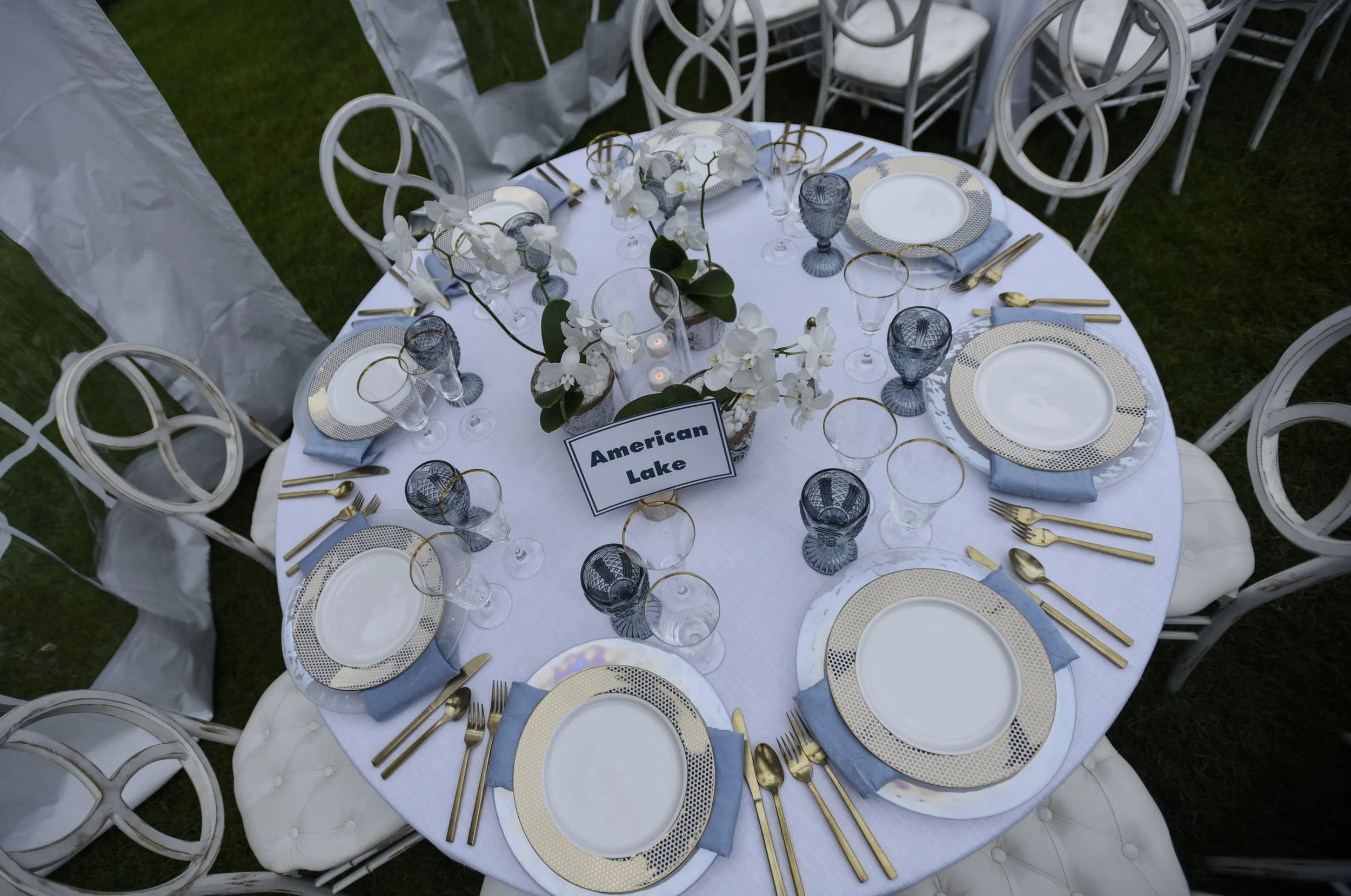 Round table set for a formal event with white and gold dishes, gold flatware, and gray napkins. A central floral arrangement features white orchids, and a sign reads "American Lake." There are glasses with gold rims and blue-tinted glasses. The table