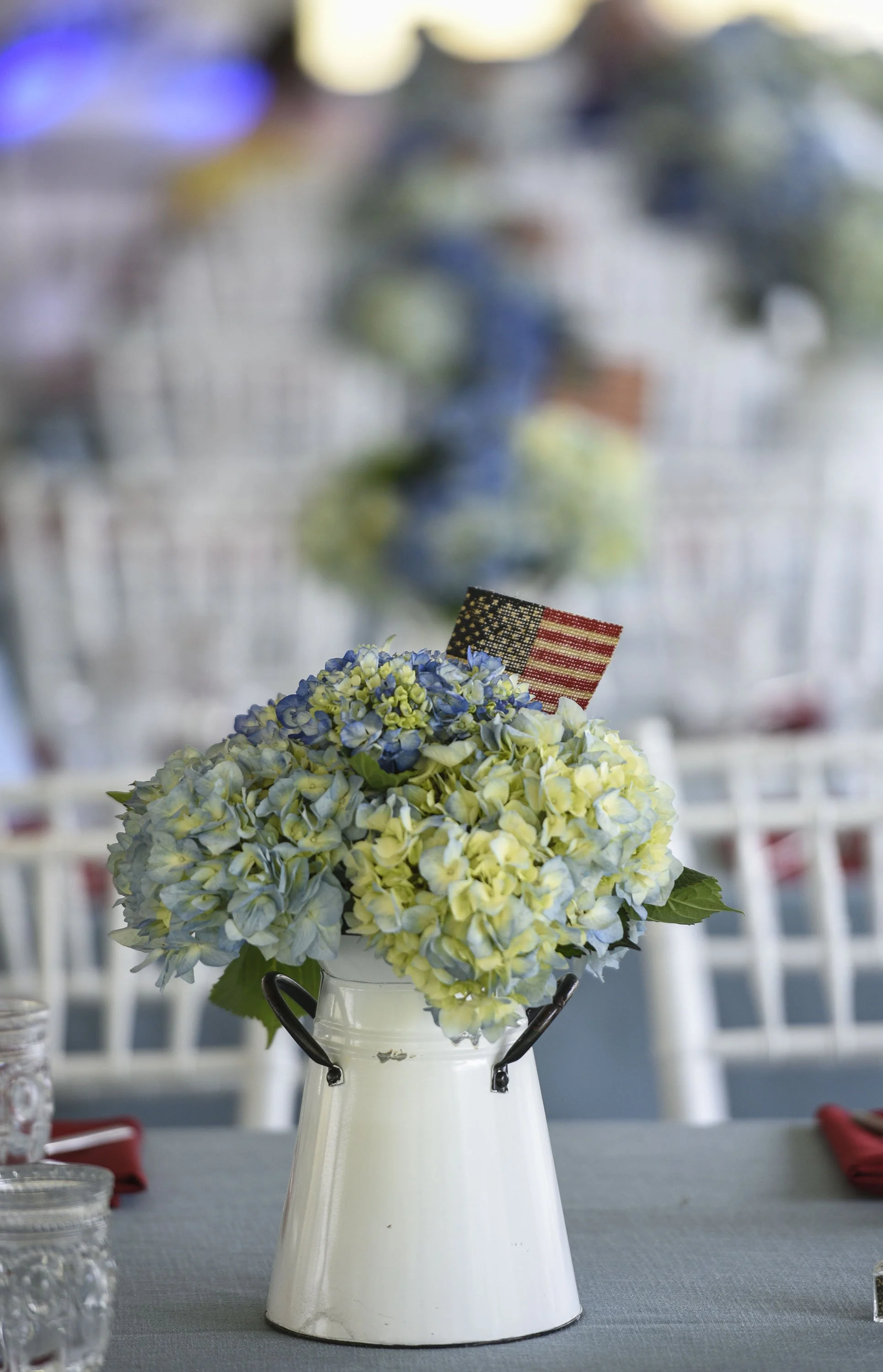 A white metal vase with handles holding a bouquet of blue and white hydrangeas, decorated with a small American flag, placed on an outdoor table with glasses and red napkins, likely at a patriotic celebration game or event.