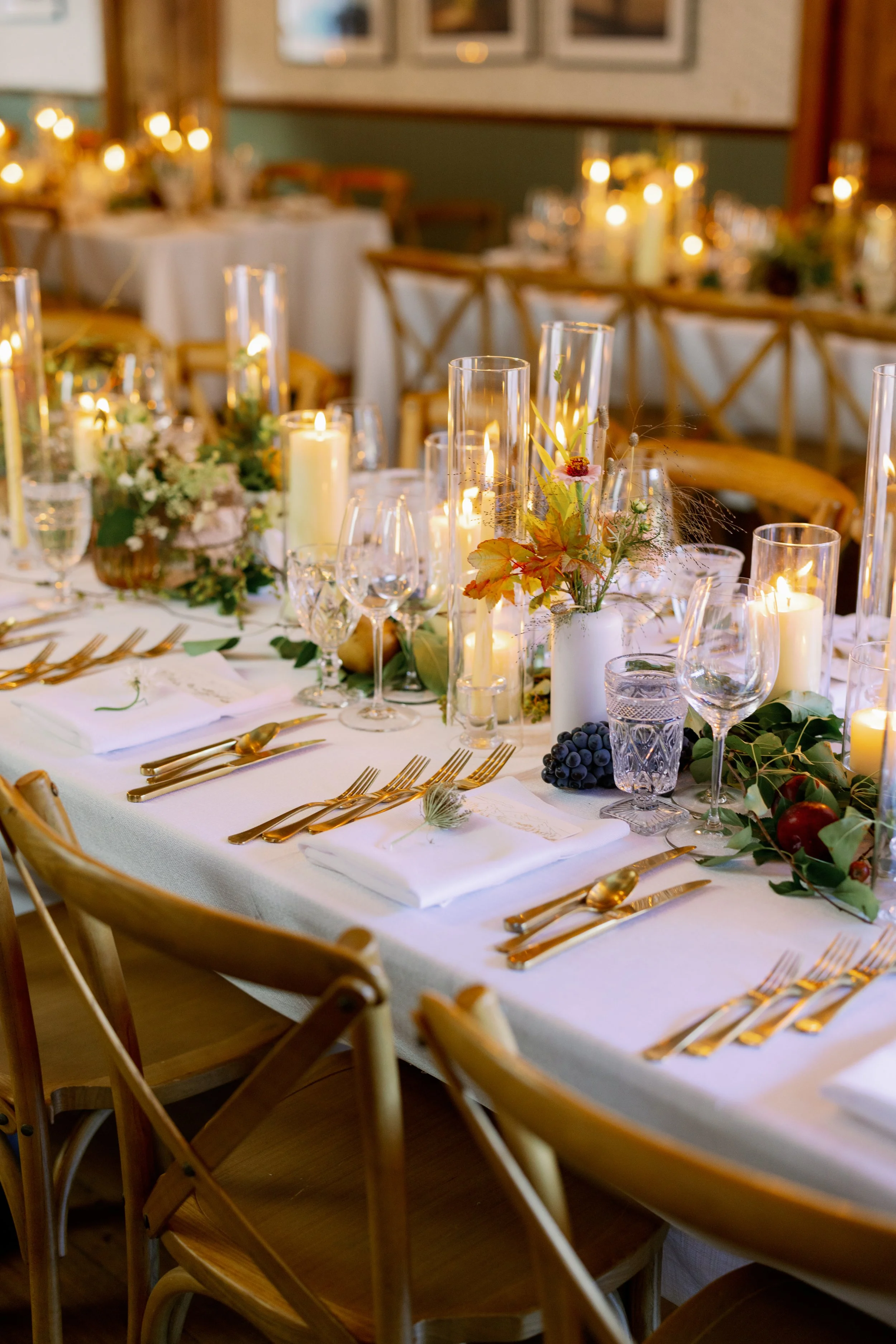 Elegant dining table set with white tablecloth, gold utensils, white napkins, and lit candles. Decor includes a centerpiece with flowers, grapes, and greenery.