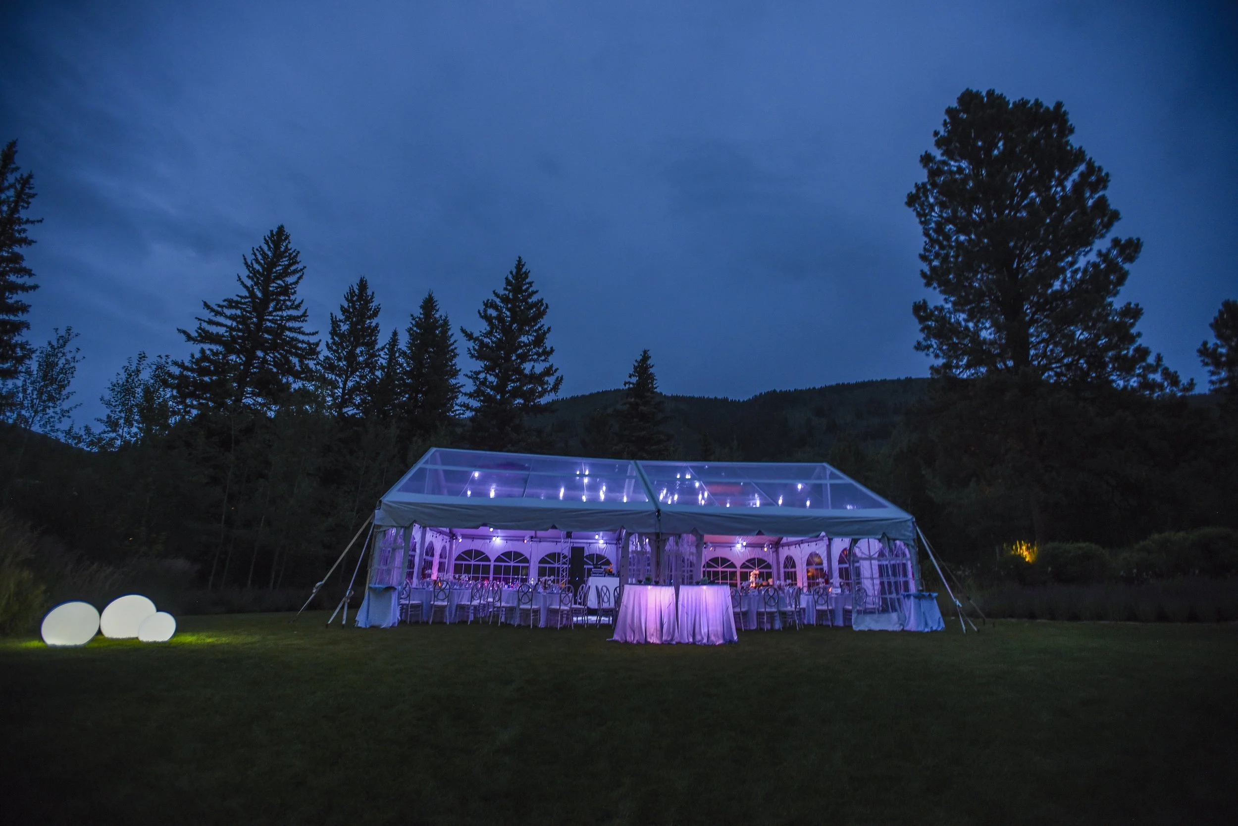 A large outdoor event tent illuminated with purple and white lights, set up on a grassy area surrounded by trees, during dusk.