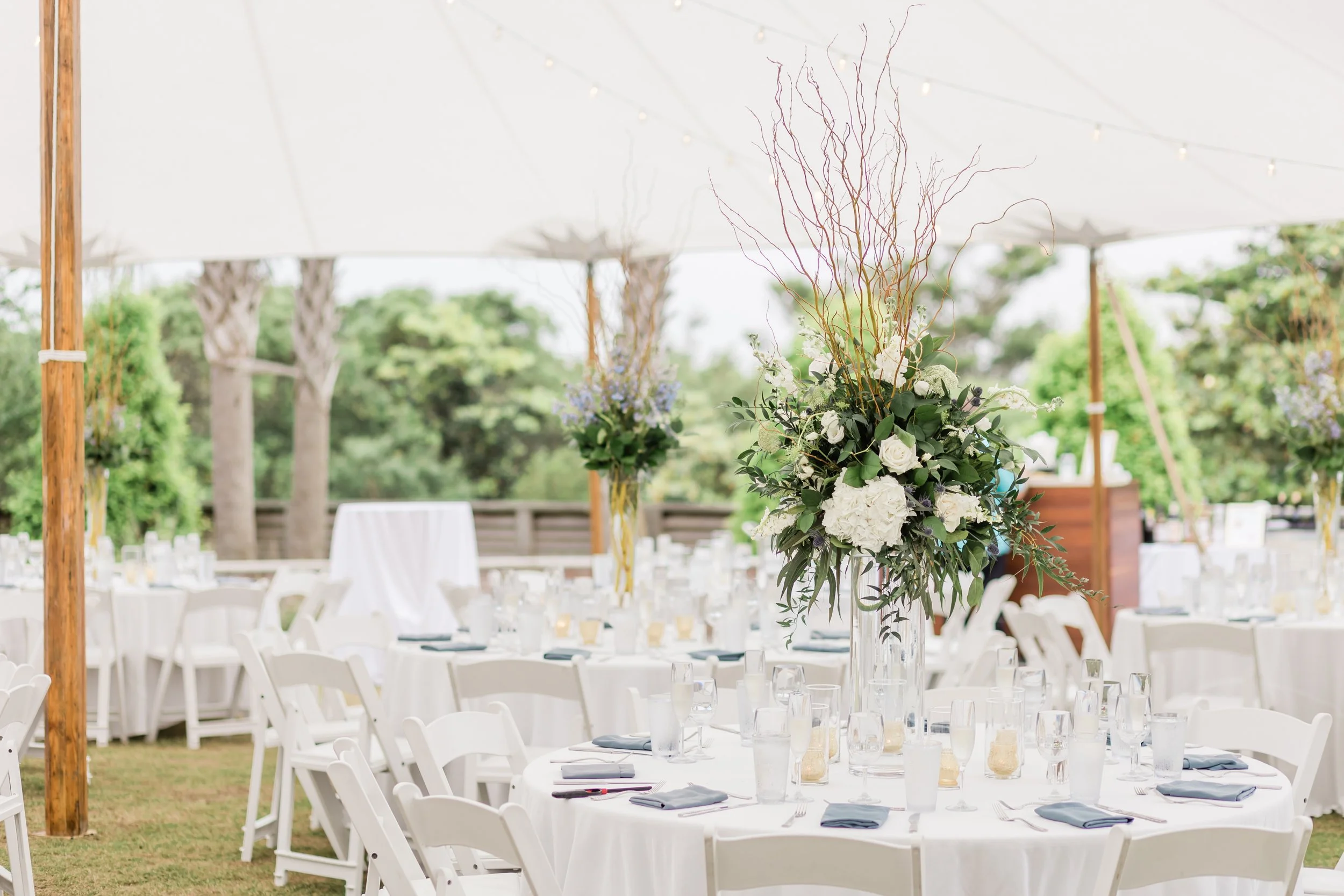 Elegant outdoor wedding reception setup with round tables draped in white cloths, each decorated with large floral centerpieces of white roses and hydrangeas, under a white tent with string lights, surrounded by greenery.