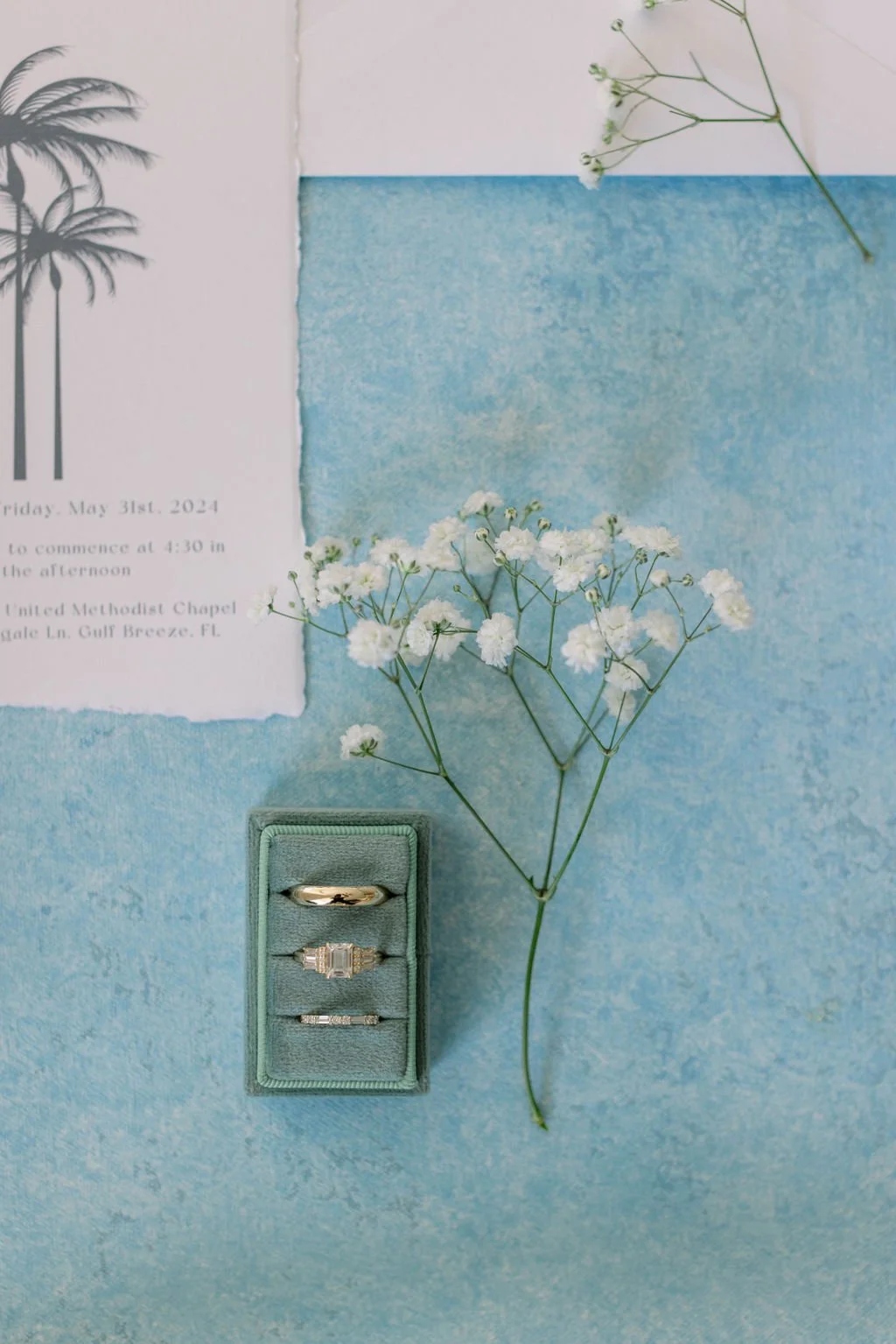 Wedding invitation with palm trees illustration, white flowers, and rings in a green ring box on a blue background.