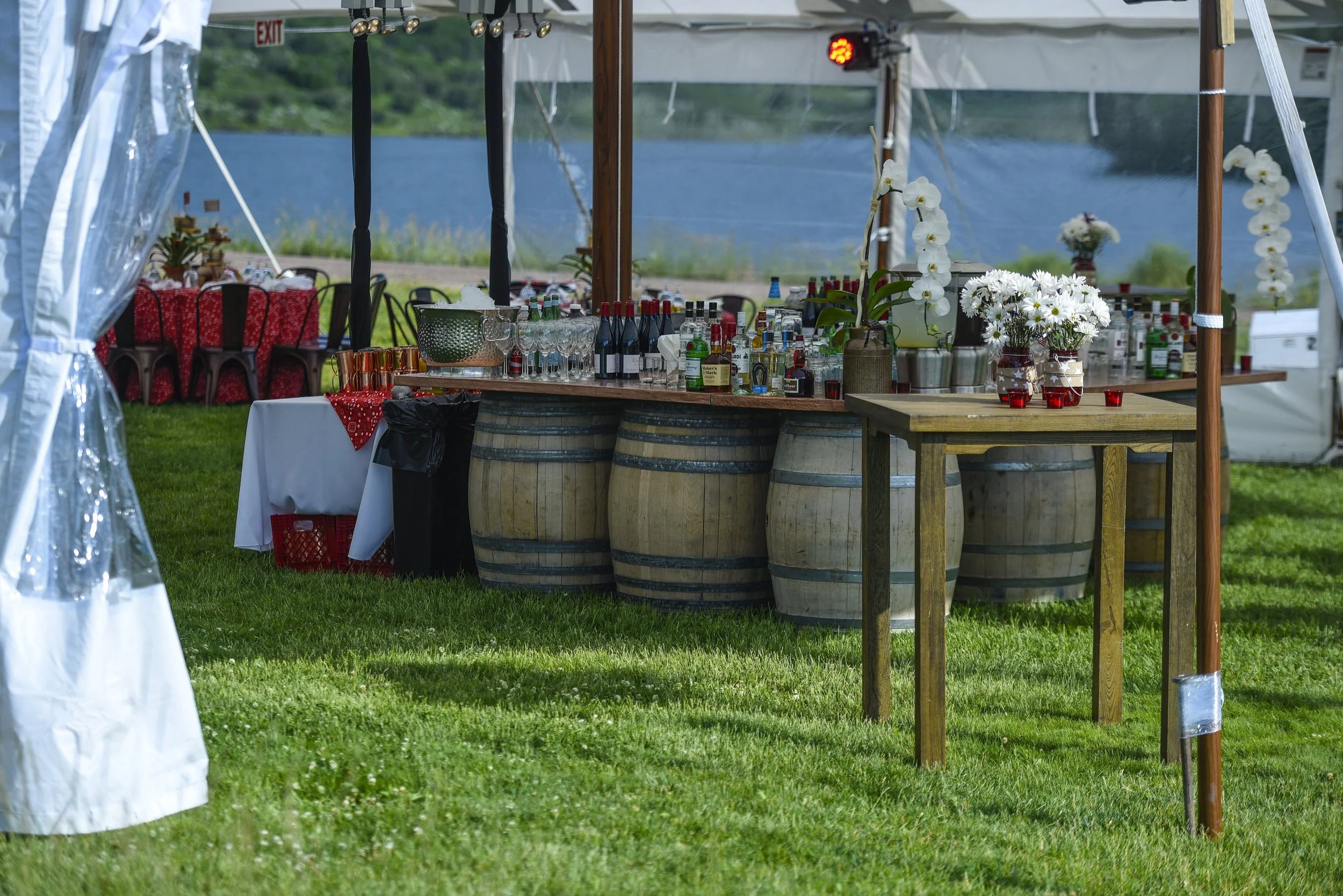 Outdoor event setup with a bar made of wine barrels, bottles, glassware, flowers, and a table, under a large white tent with a grassy area and lake in the background.