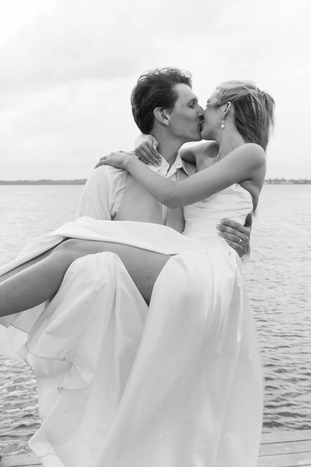 A black and white photo of a bride and groom kissing, with the groom holding the bride, near a body of water.