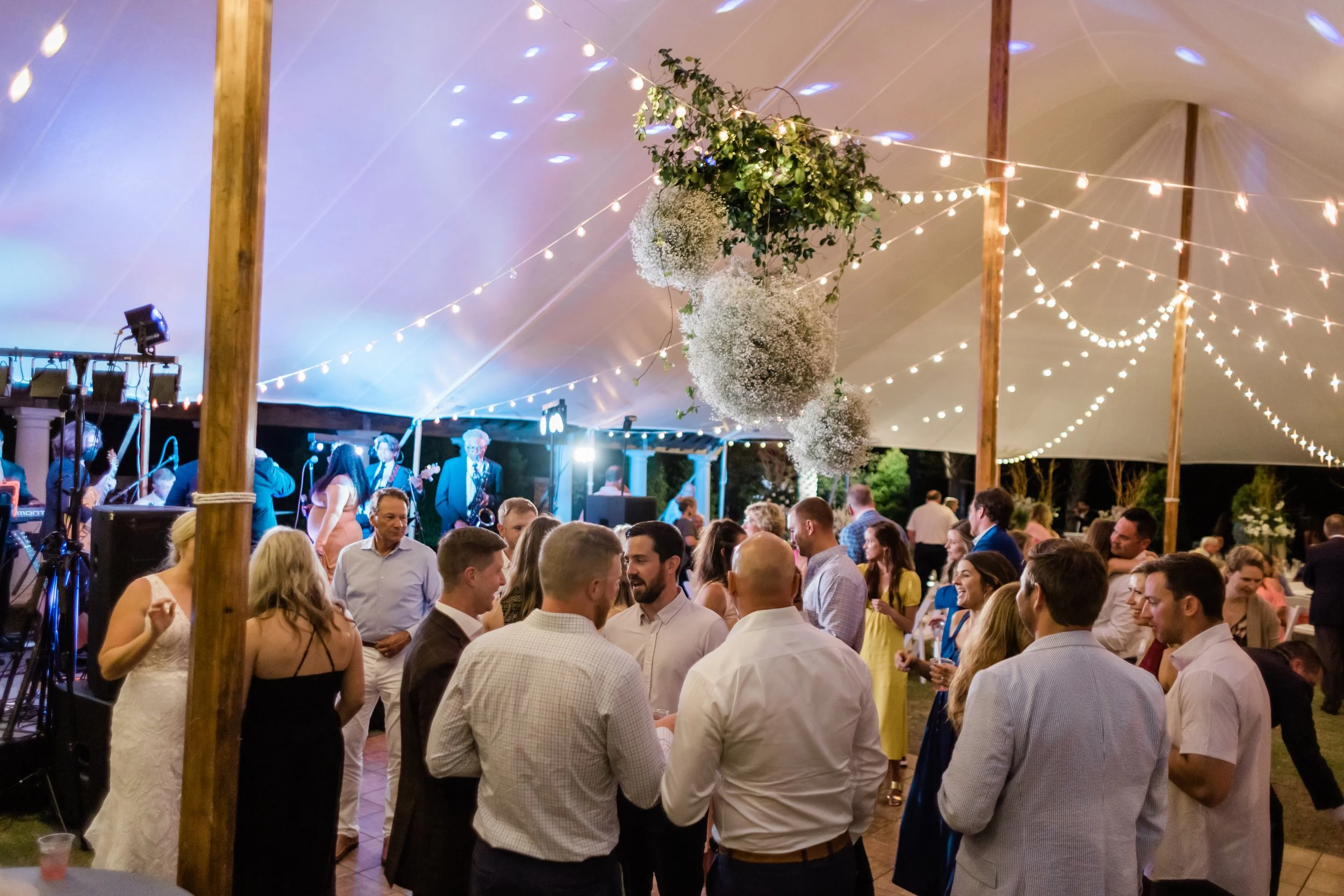 Guests dancing and socializing at a wedding reception under a large tent decorated with hanging floral arrangements and string lights, with a band playing on a stage in the background.