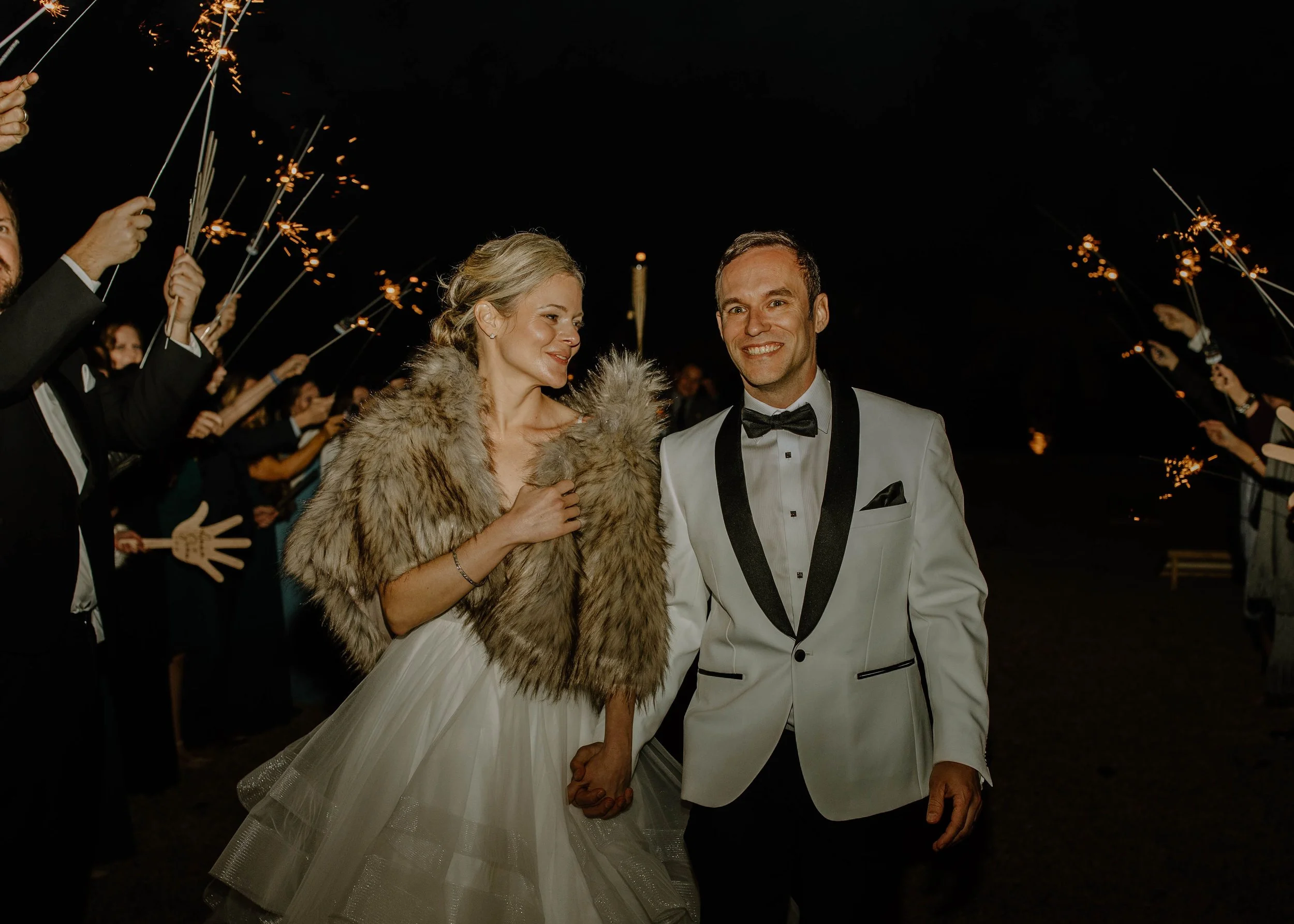 A newlywed couple holding hands and smiling, surrounded by people holding sparklers at night during a wedding celebration.
