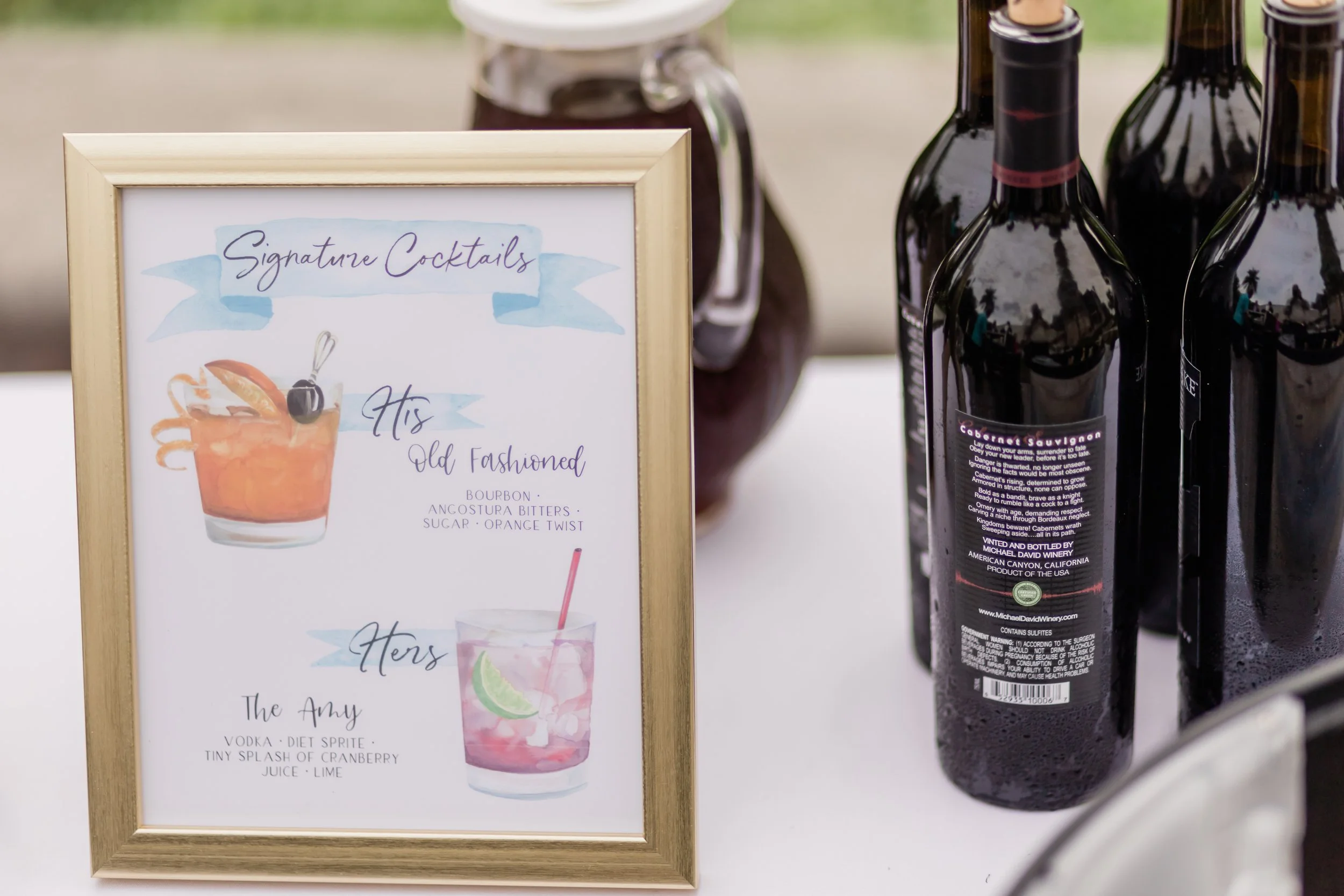 A menu board displaying signature cocktails, featuring two drinks labeled 'His Old Fashioned' with bourbon, Angostura bitters, sugar, orange twist, and 'Hers The Amy' with vodka, diet Sprite, cranberry juice, and lime, with bottles of wine or beer be