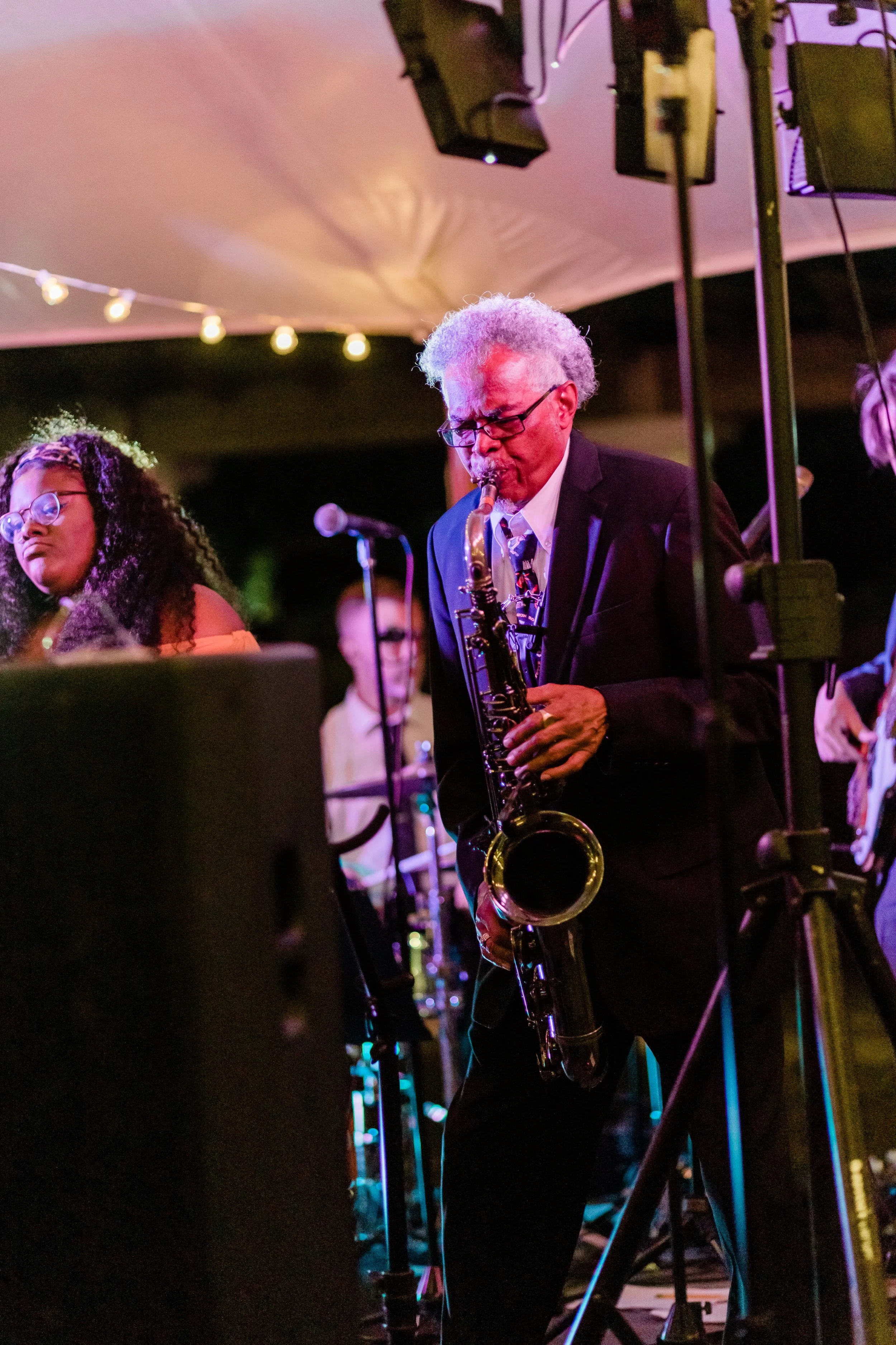 An elderly man with gray hair and glasses playing a saxophone during a live jazz performance at night, under a tent with string lights.