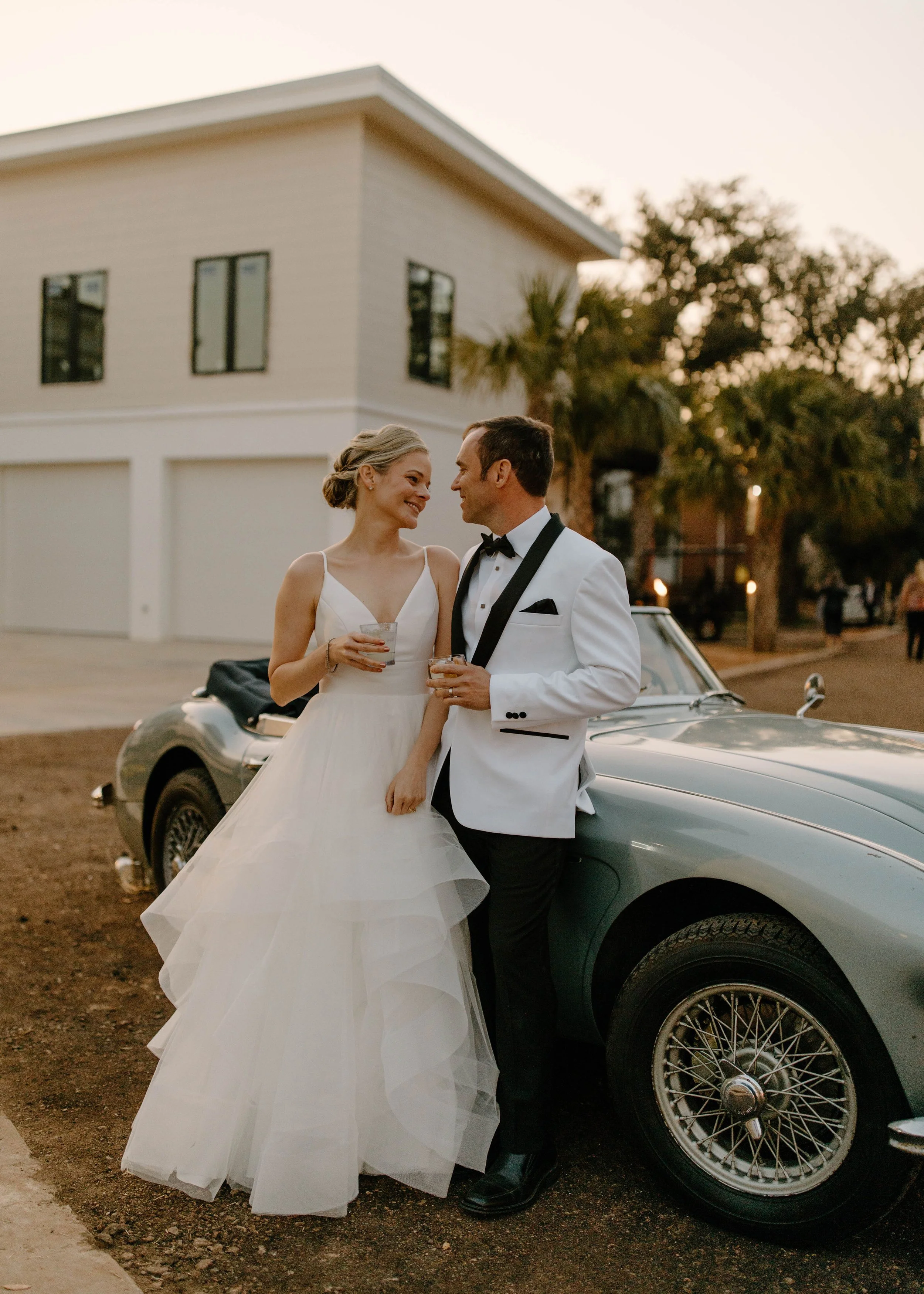 A bride and groom in wedding attire standing next to a vintage silver car outside during sunset, smiling and holding drinks.