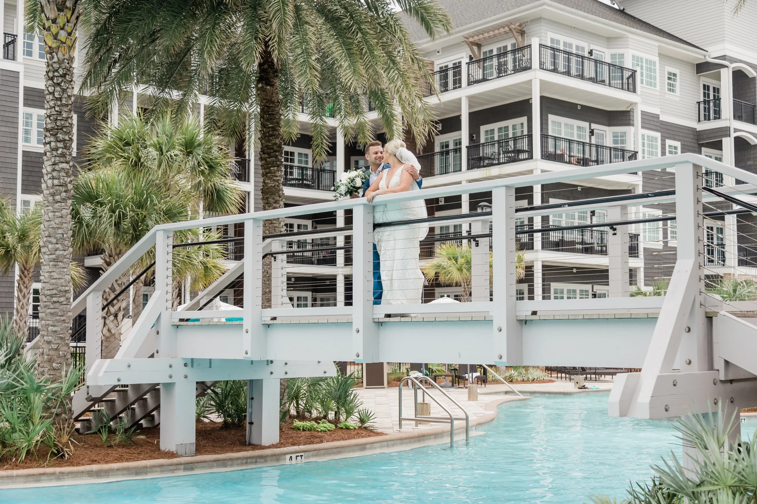 A newlywed couple stands on a bridge over a swimming pool at a resort, sharing a kiss. The bride is holding a bouquet of flowers, and the groom is smiling. The background features multi-story modern hotel buildings with balconies and palm trees.