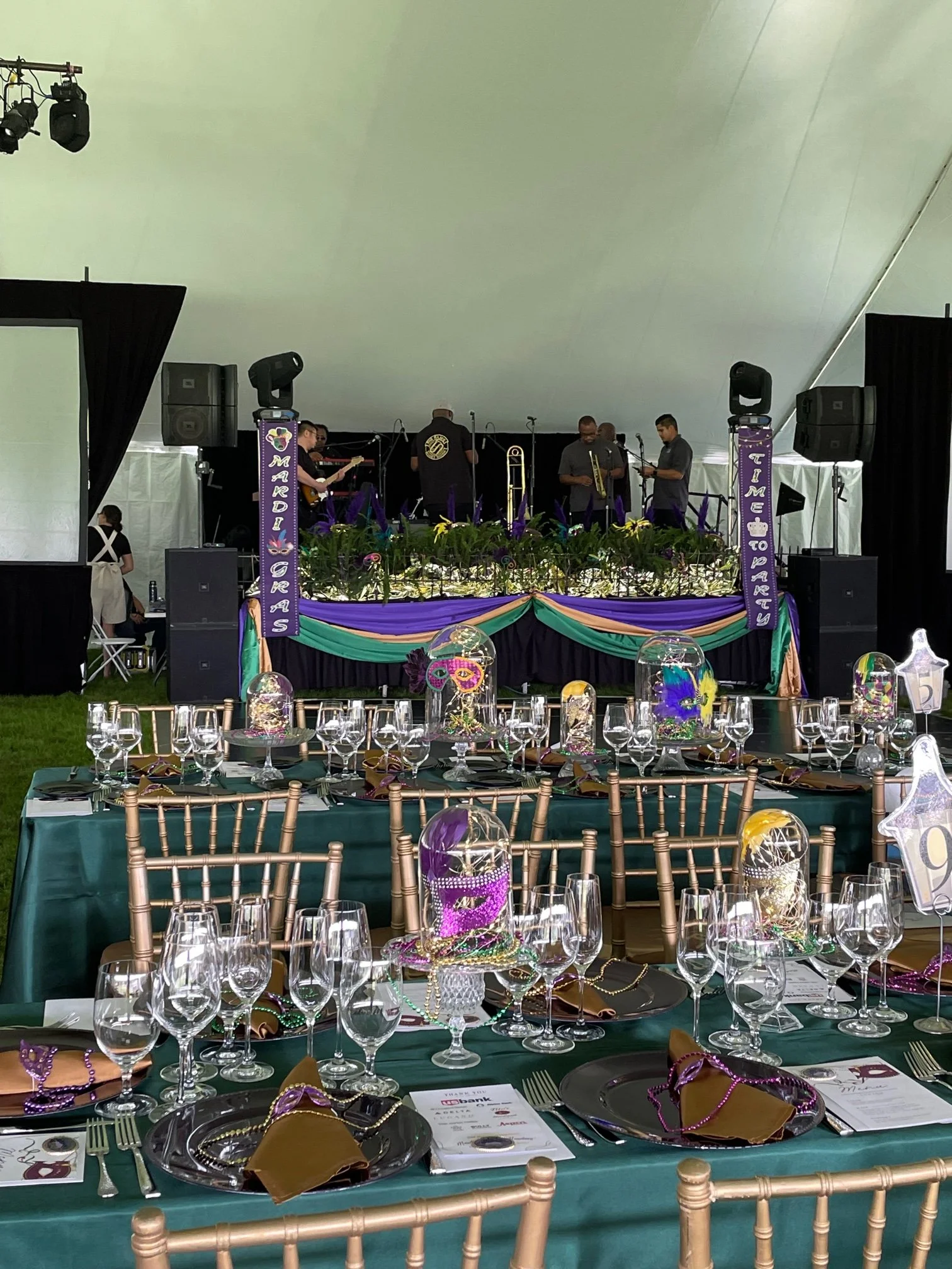 Decorated event tent with tables set for a celebration, purple and gold Mardi Gras themed decorations, a stage with a live band, and chairs arranged around the tables.