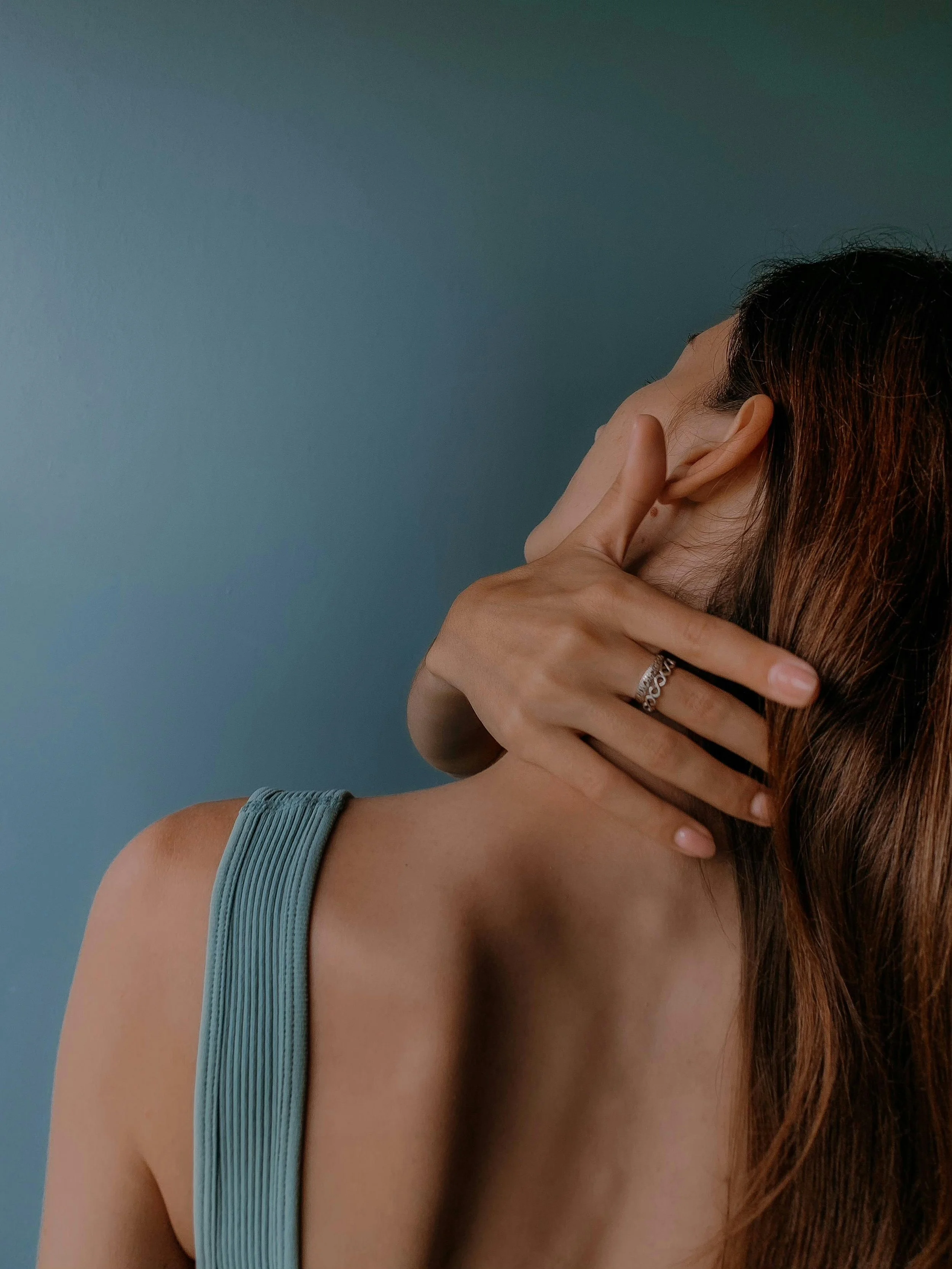 A woman with long brown hair, wearing a blue tank top, holding her hand near her ear, with a light blue wall in the background.