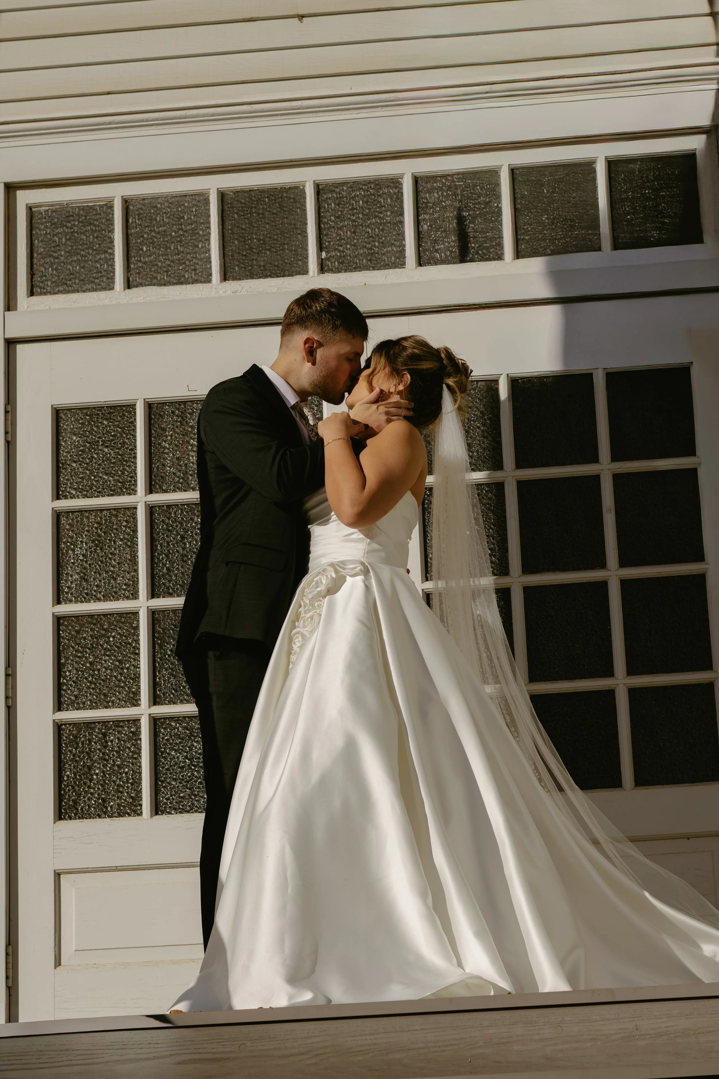 A bride and groom share a kiss in front of a large garage door, with the bride wearing a white wedding gown and veil, and the groom in a black suit.