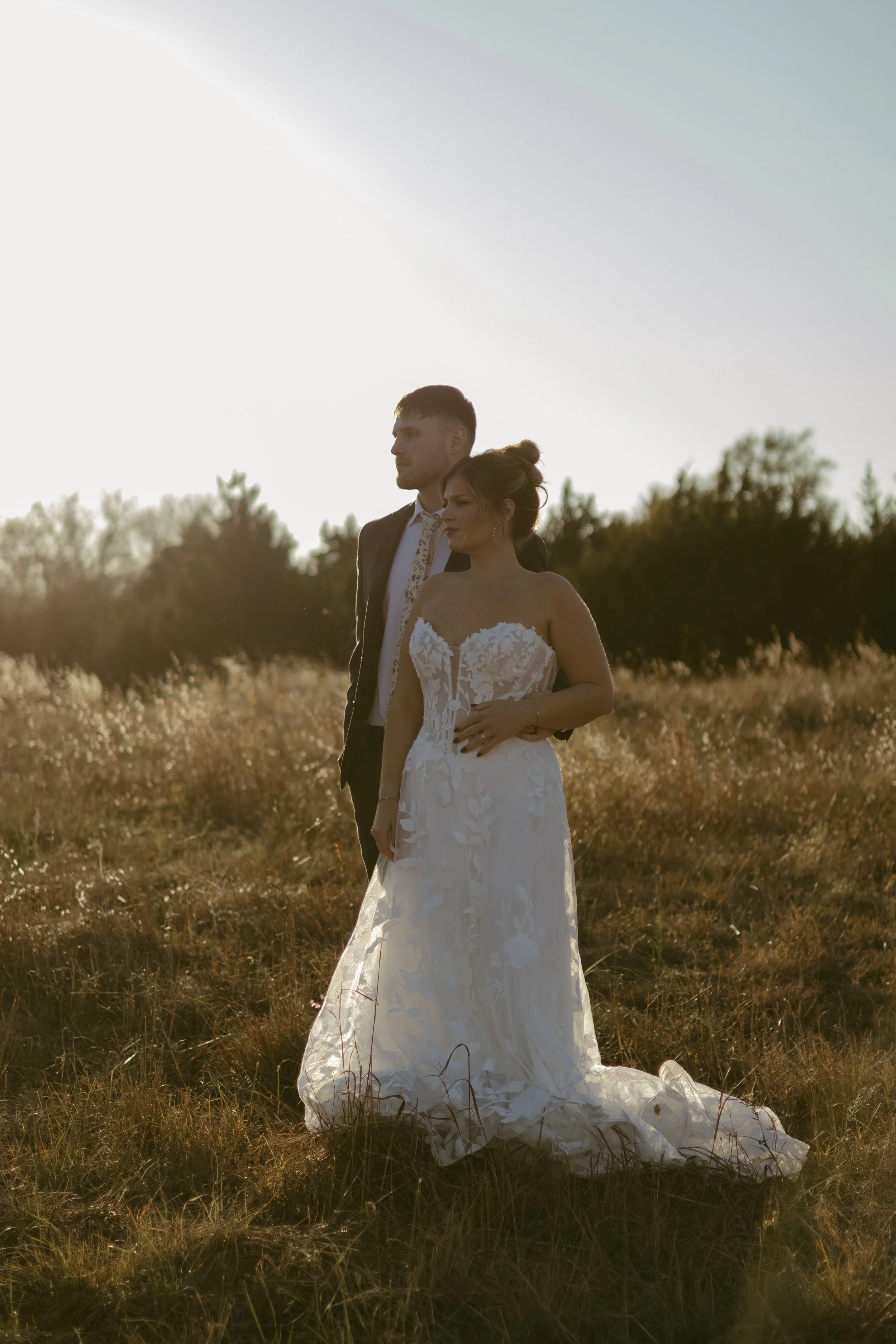 A bride and groom standing in a field during sunset, with the bride in a white wedding dress and the groom in a dark suit.