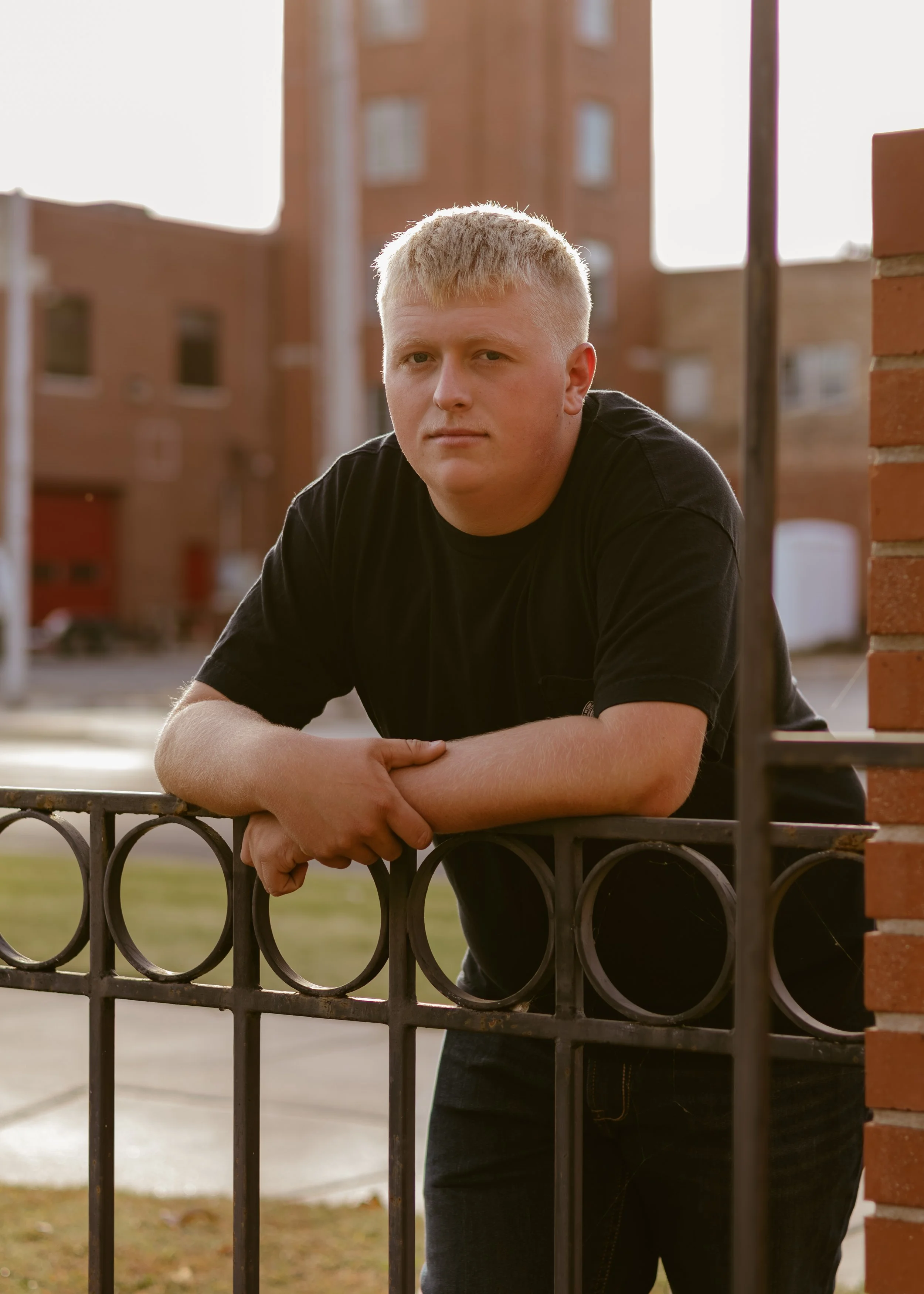 A young man with blonde hair and a serious expression leaning on a black iron fence outdoors in front of brick buildings during the daytime.