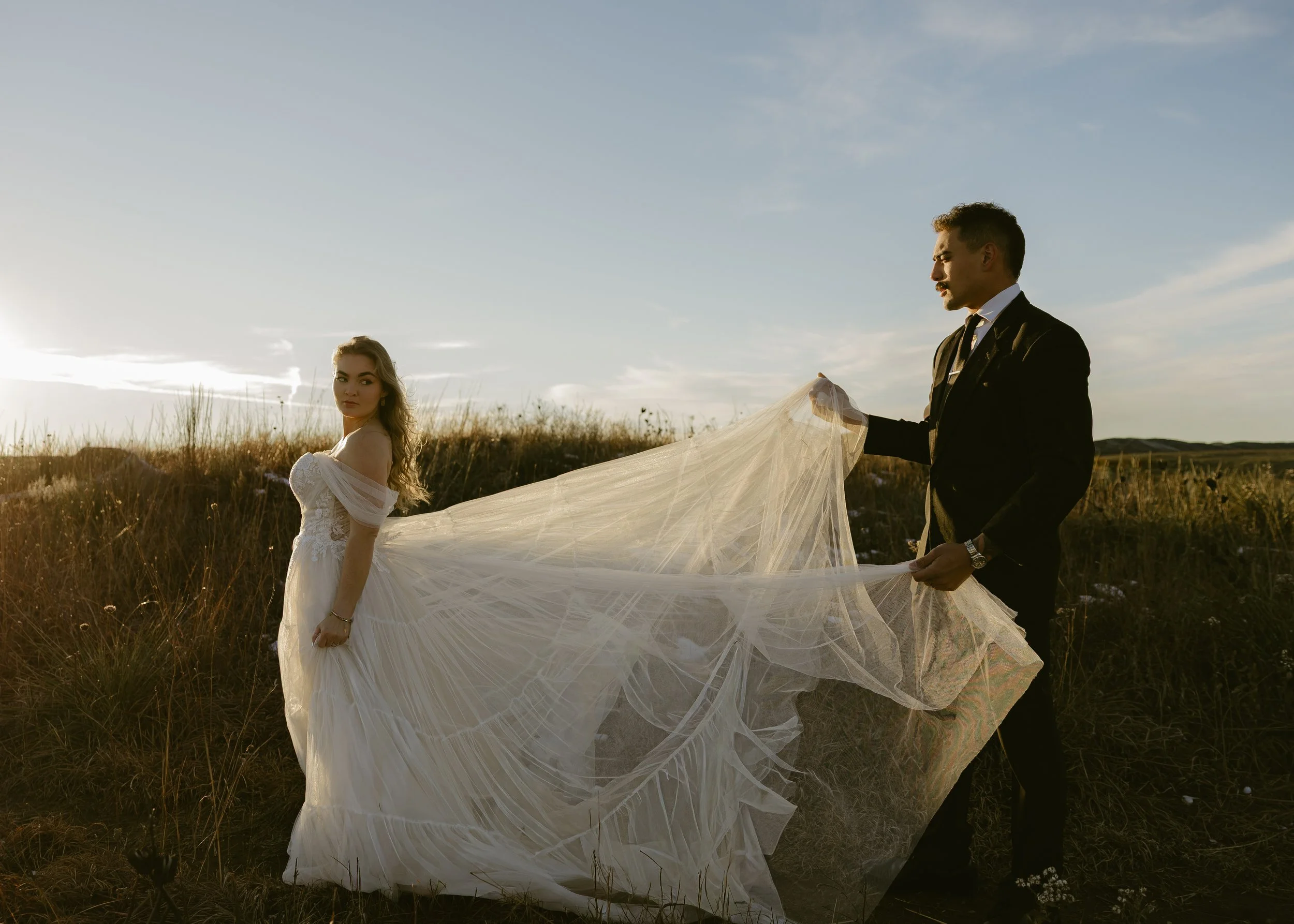 A man in a black suit holding a bridal veil, standing in a field with a woman in a white wedding gown, both during sunset.