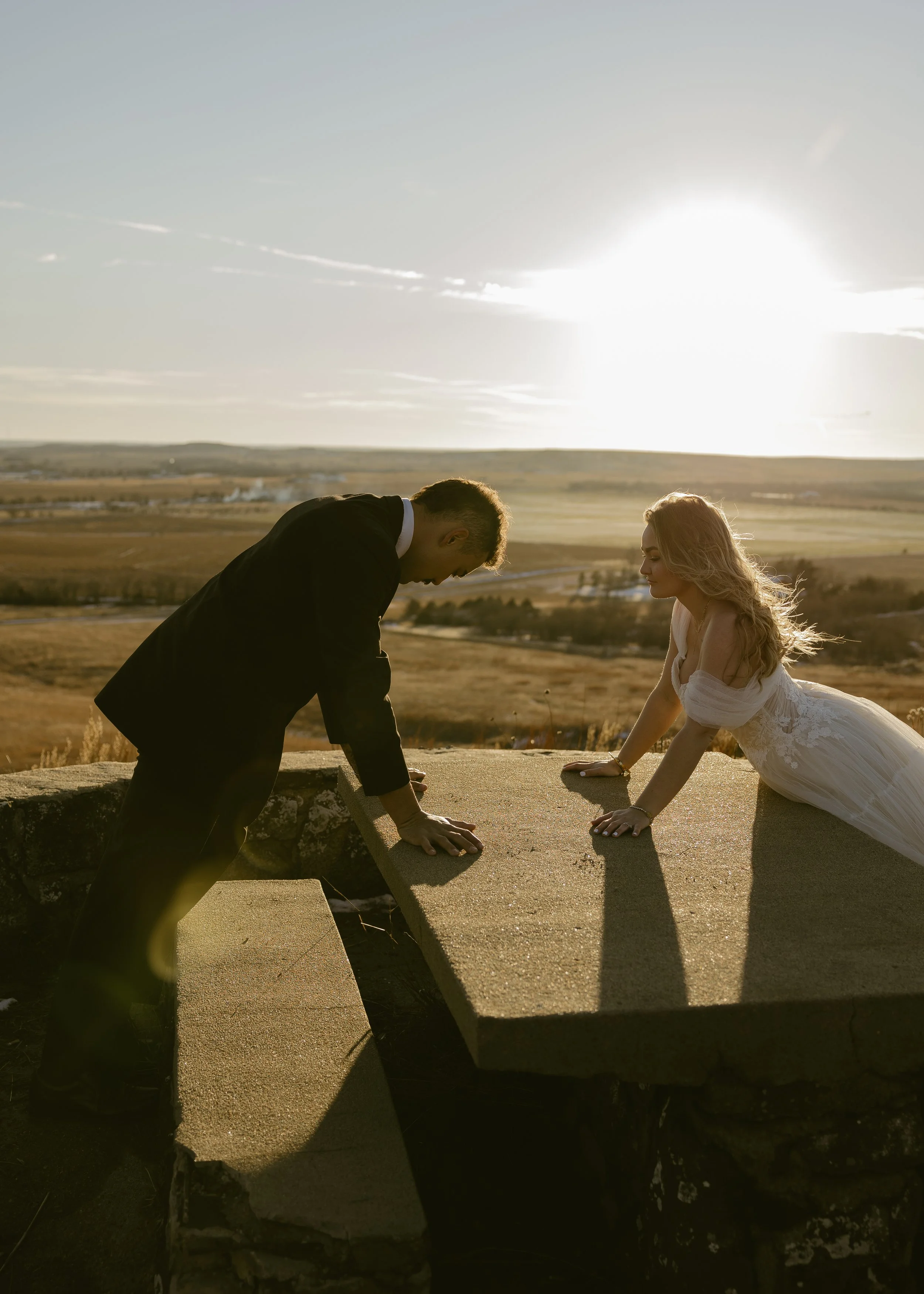 A couple dressed in wedding attire, a man in a suit and a woman in a wedding dress, doing push-ups on a stone surface outdoors during sunset with a vast open landscape in the background.