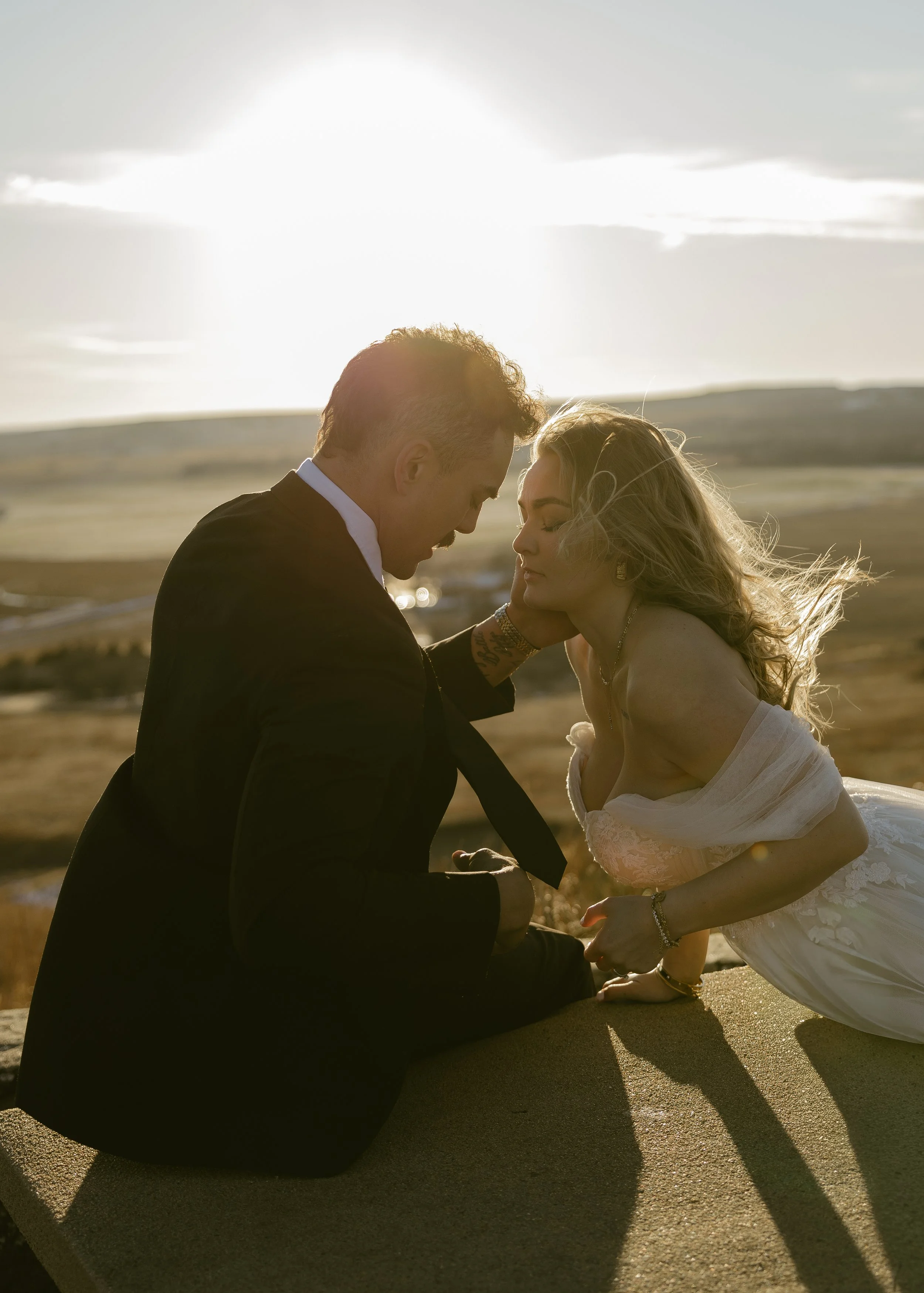 A man in a black suit and a woman in a white dress are kneeling on a sandy surface outdoors during sunset, touching foreheads closely amid a scenic landscape with open fields and cloudy sky.