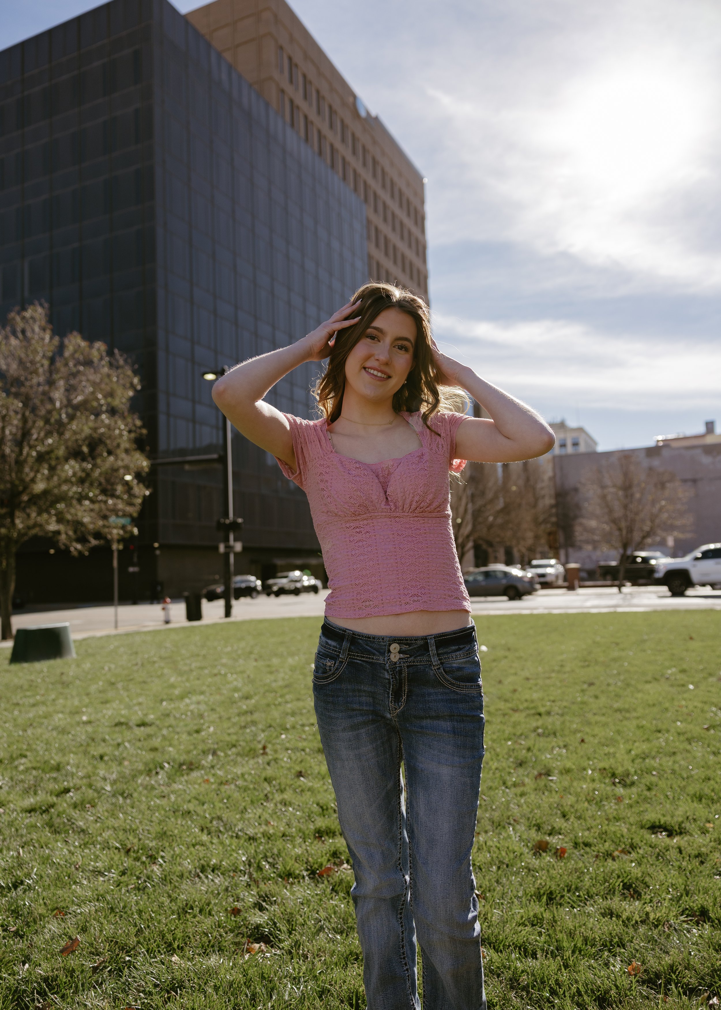 A young woman standing outdoors on a grassy area, smiling, with her hands on her head, wearing a pink short-sleeved top and jeans, with a large building and cars in the background on a sunny day.