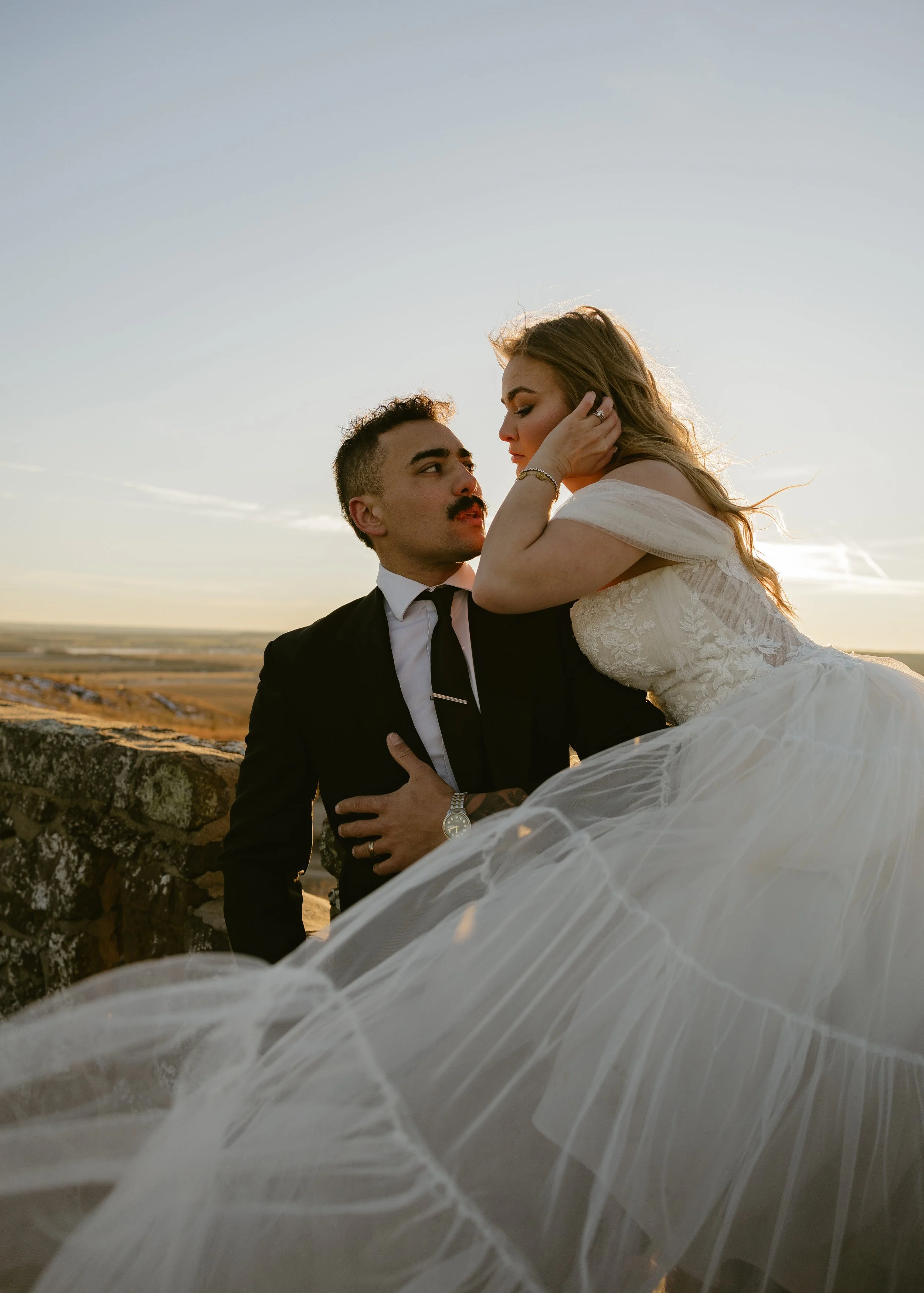 A bride and groom sharing an intimate moment outdoors during sunset, with the bride sitting on a stone ledge and the groom looking up at her.