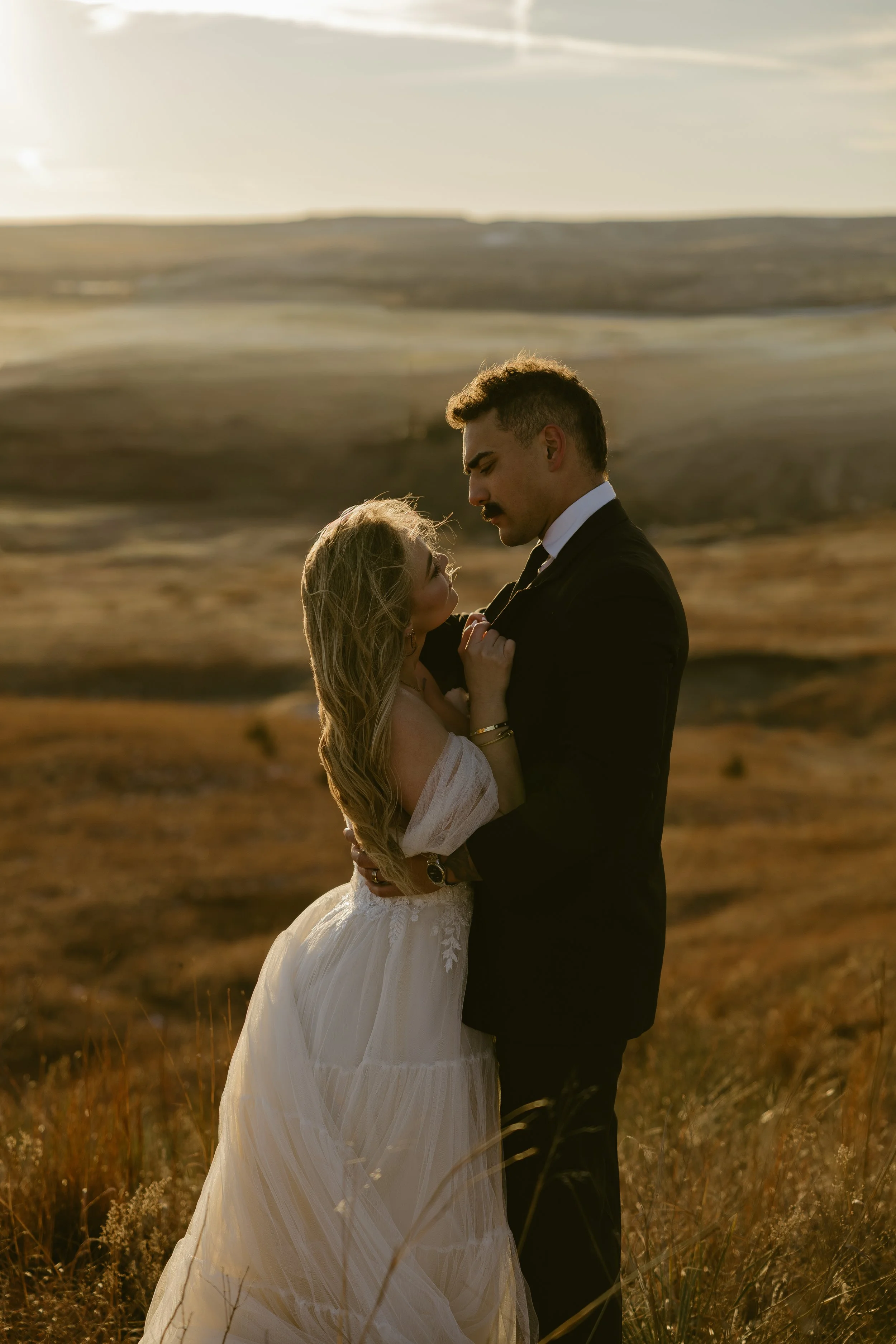A couple embraces in a grassy field during sunset, with the woman in a white dress and the man in a black suit.