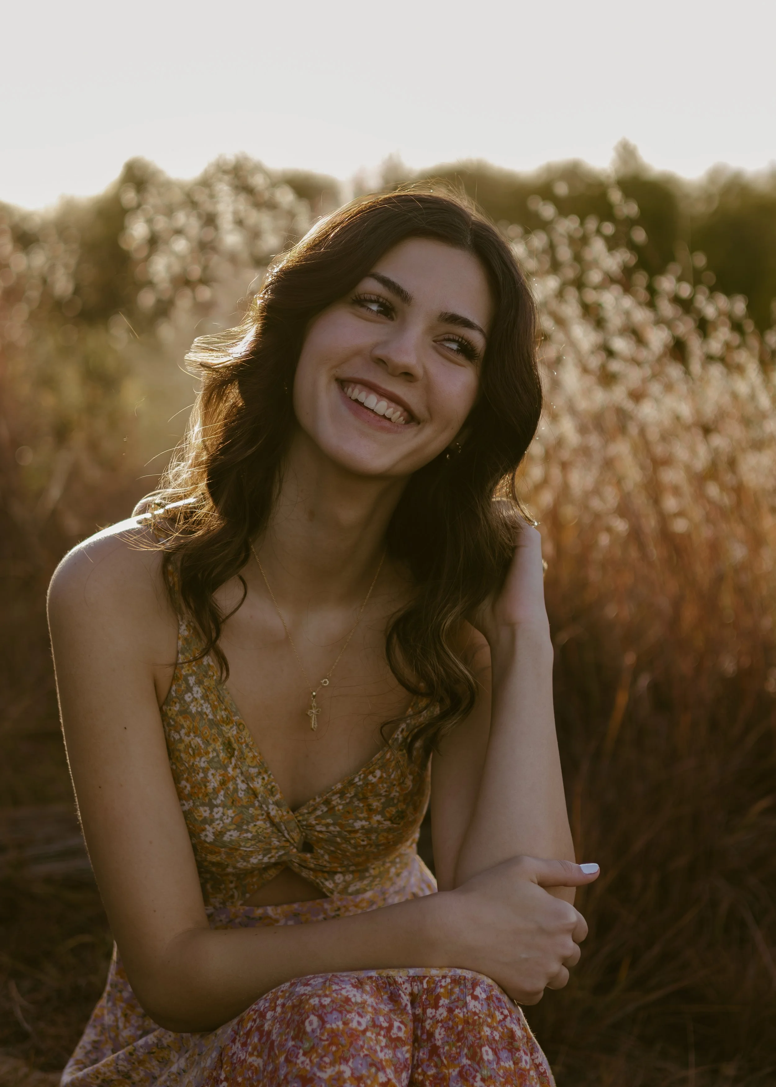 A young woman with wavy brown hair smiling outdoors during sunset, wearing a floral dress and a gold cross necklace, sitting in a field with tall grass and trees in the background.