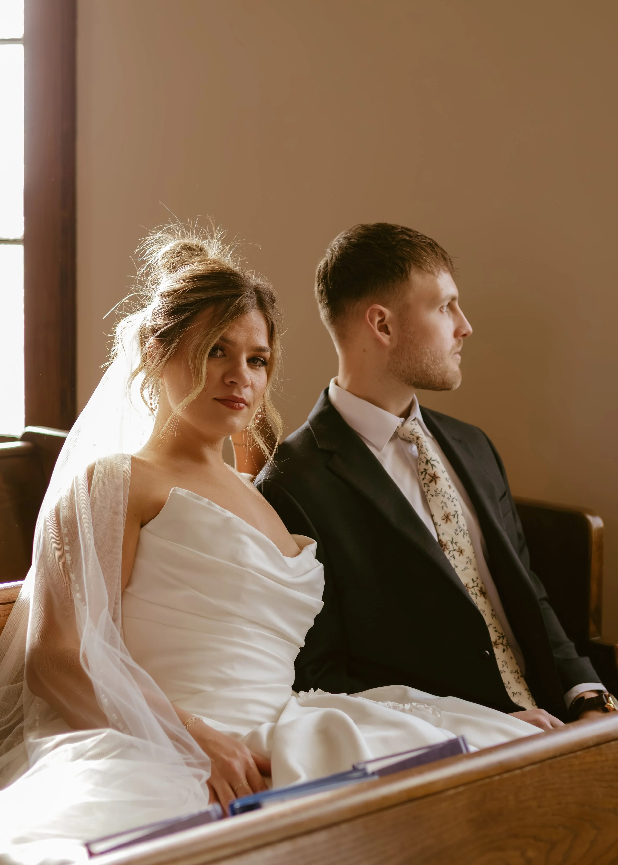 A bride and groom sitting in a church, with the bride looking at the camera and the groom looking away.