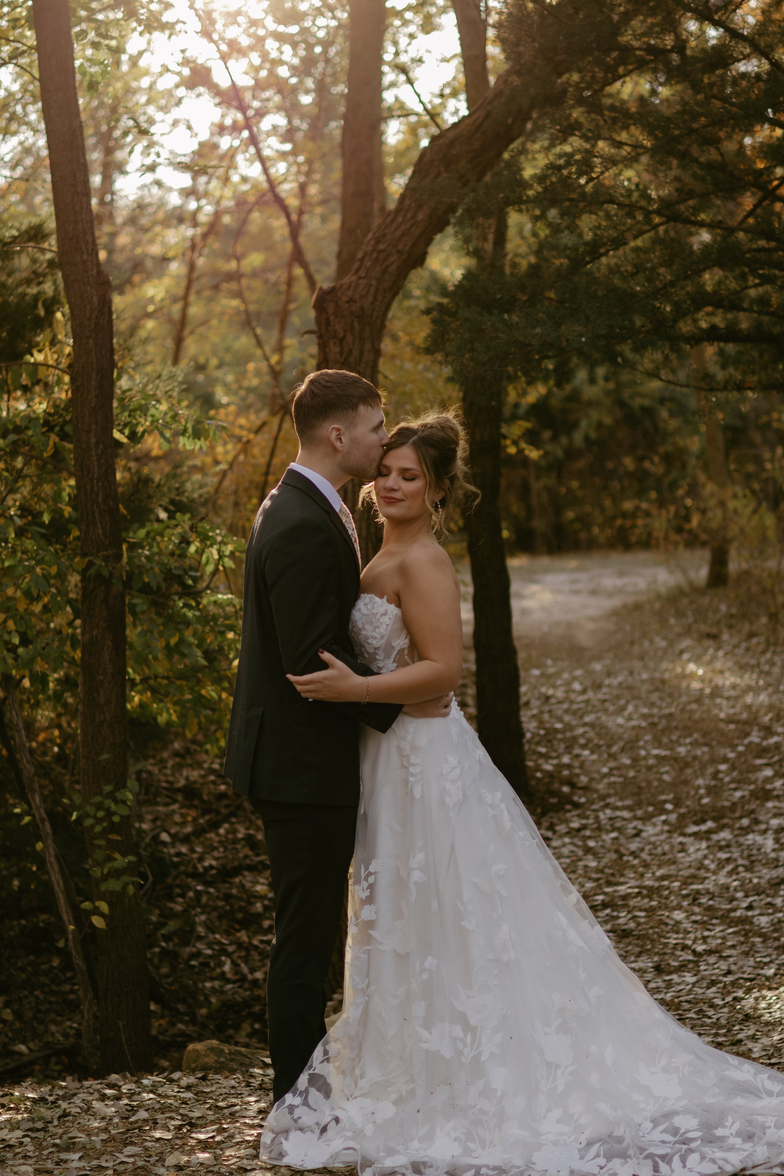 A bride and groom embracing and sharing an intimate moment in a forested area during sunset, with the groom gently kissing the bride's forehead, both dressed in wedding attire.