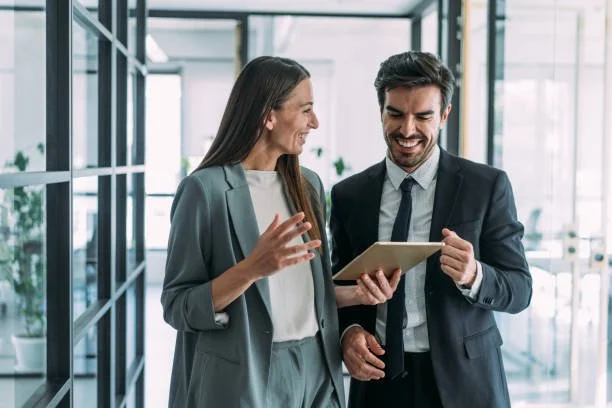 Businesswoman and businessman smiling while looking at a tablet in a modern office.